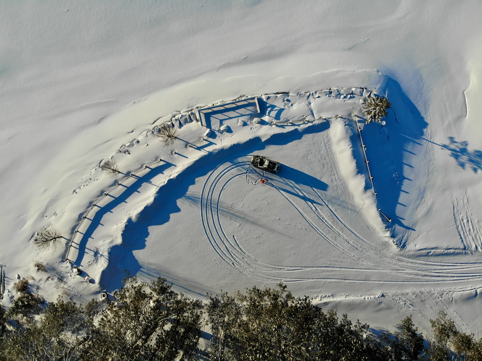 Top-down aerial view of a vehicle turning through deep snow, its tracks curving beside a fence