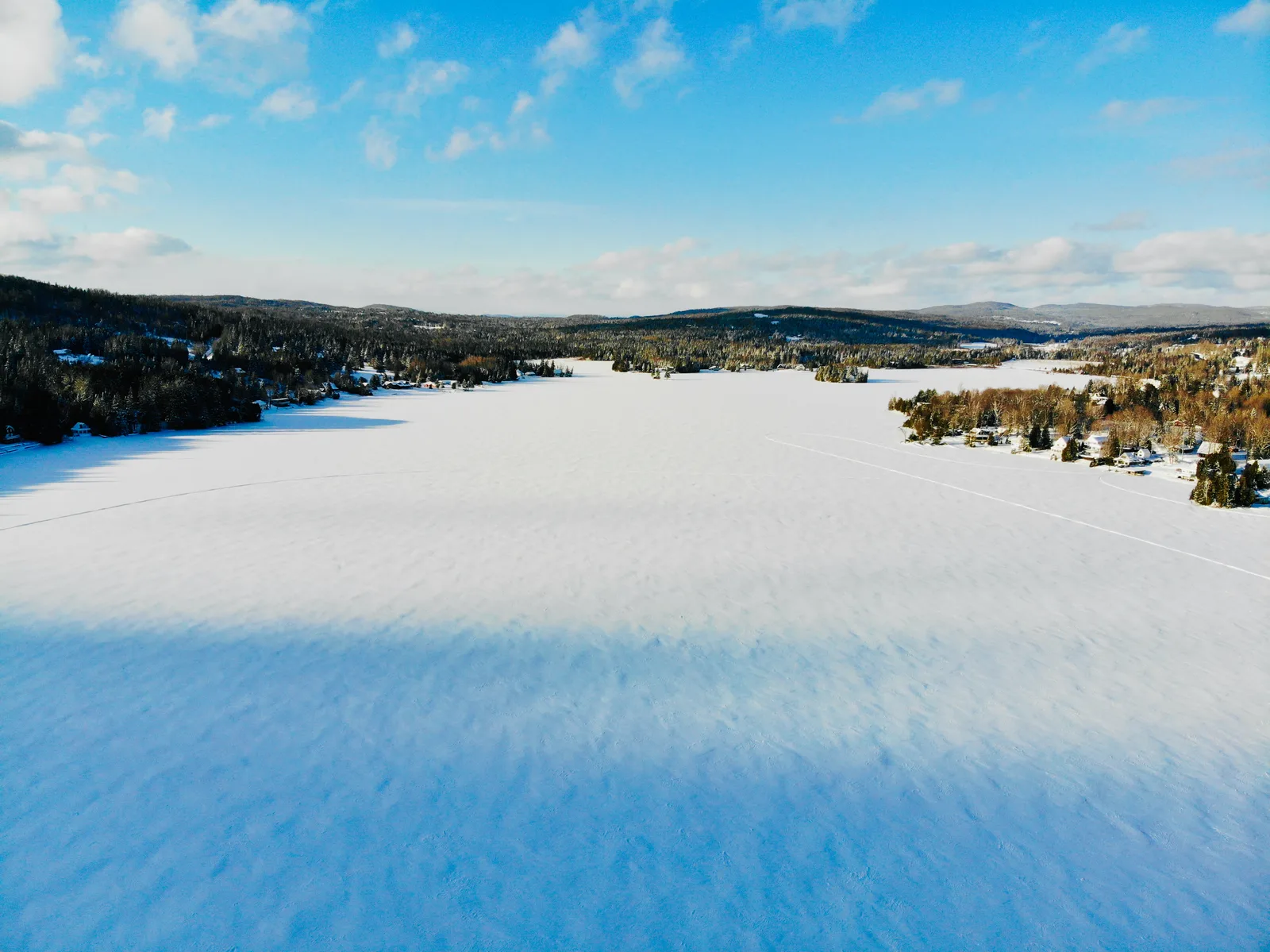Aerial view across a broad frozen lake toward wooded hills beneath a bright blue sky