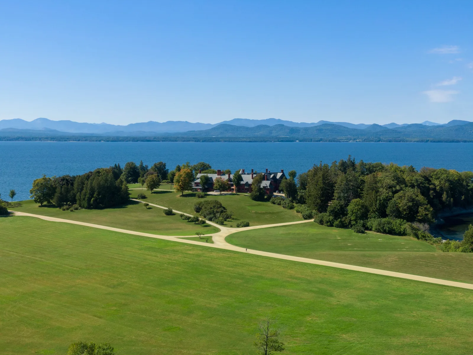 Aerial view across a lakeside lawn and tree-lined shore toward a mountain range