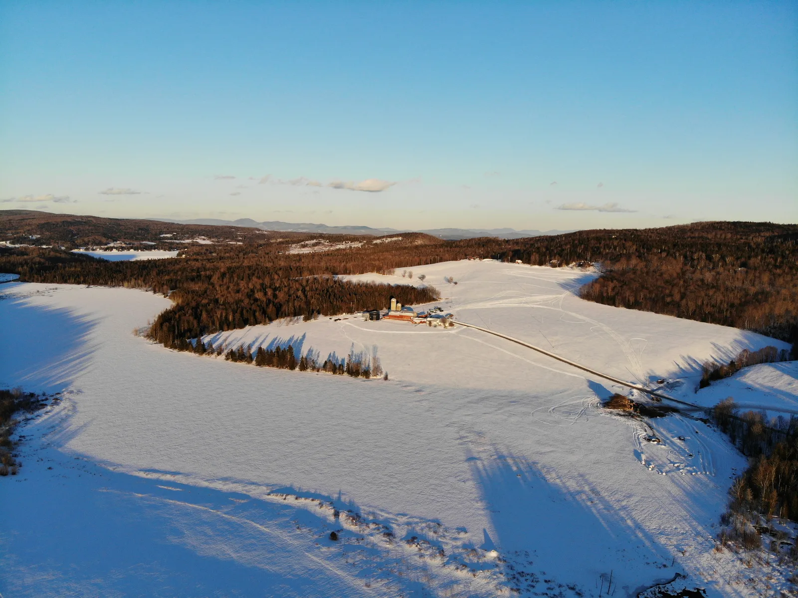 Snow-covered farm fields, a narrow road, and wooded hills in warm evening light