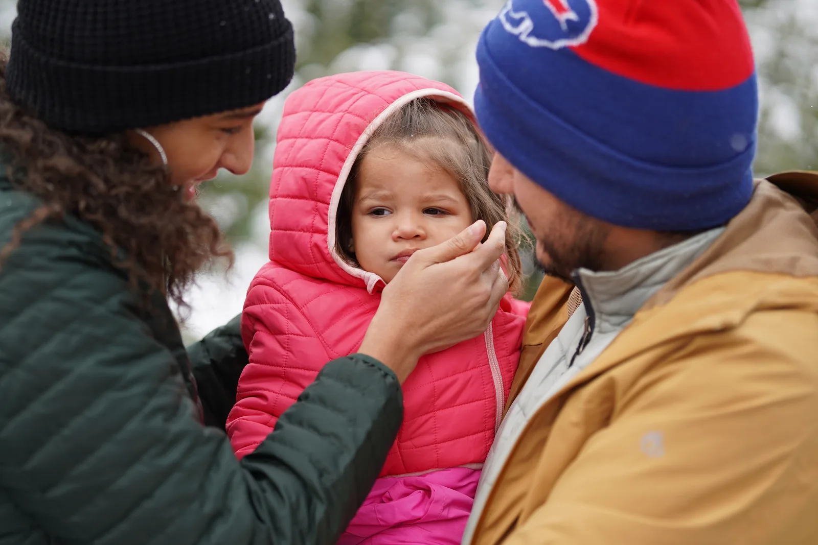 Three people in winter coats, one in a pink hooded jacket, standing close in a snowy tree farm.