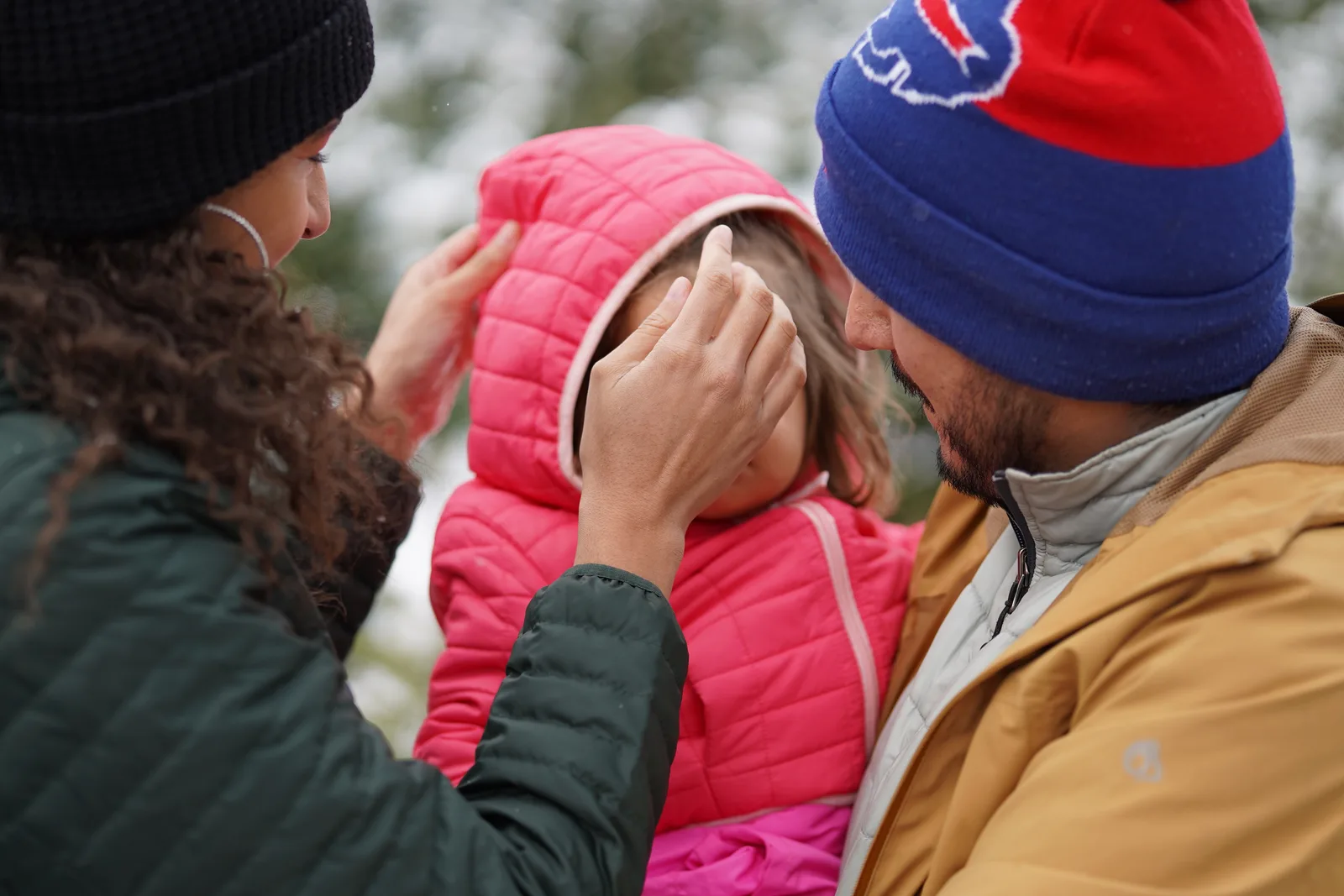 Two adults adjusting a child’s pink hooded jacket in a snowy outdoor setting.
