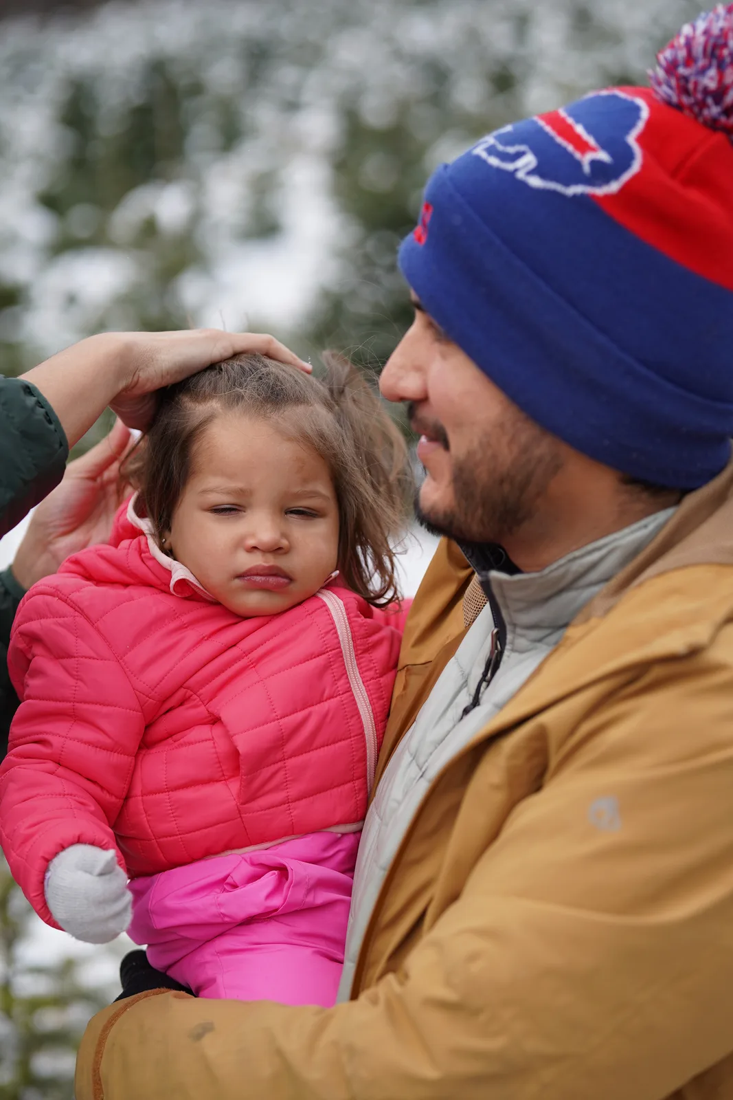 Adult in a blue and red beanie touching a child’s head in a snowy tree farm.
