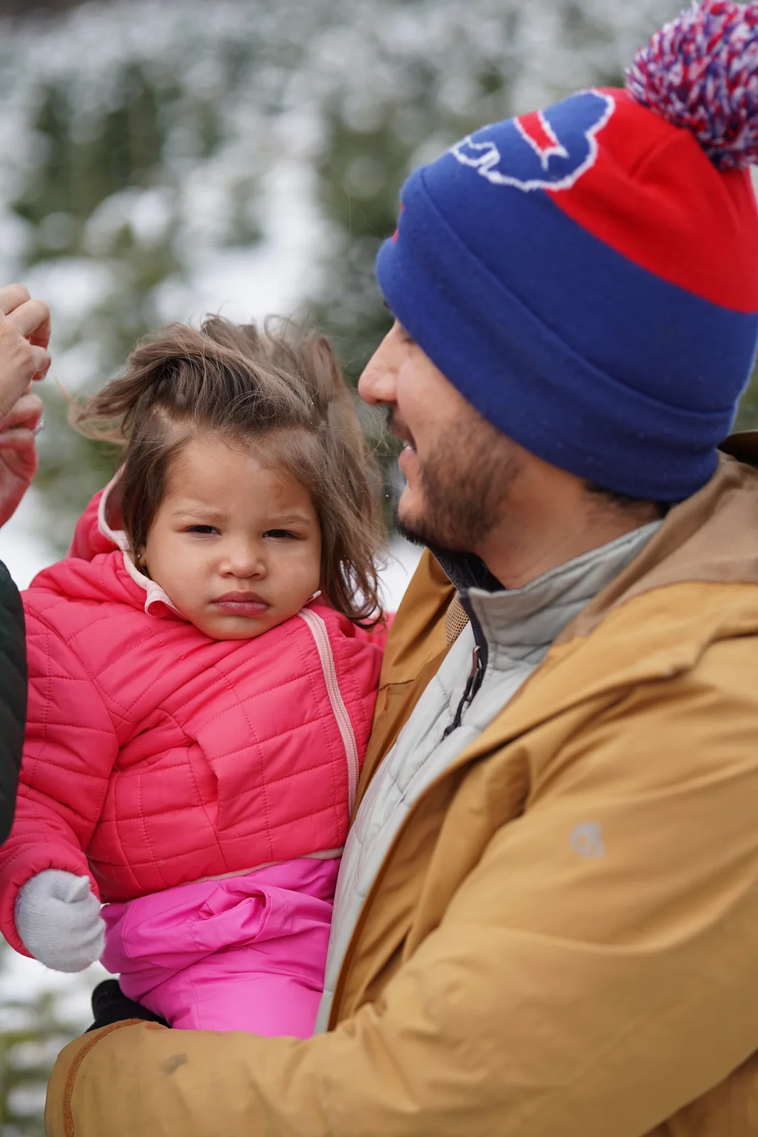 Adult in a blue and red beanie holding a child in a pink jacket among snow-covered trees.