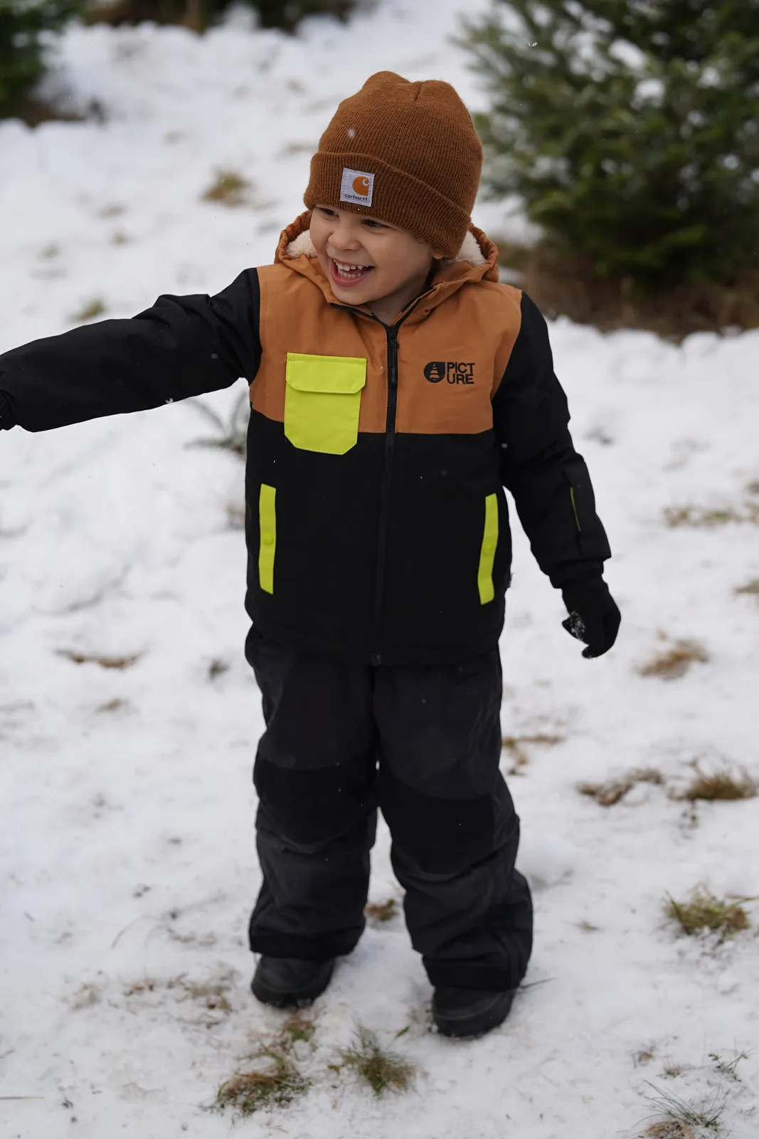 Child in a brown beanie and black jacket with yellow accents standing in snow.