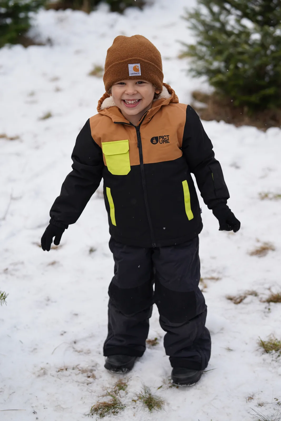Child in a brown beanie and black jacket with yellow accents smiling in snow.