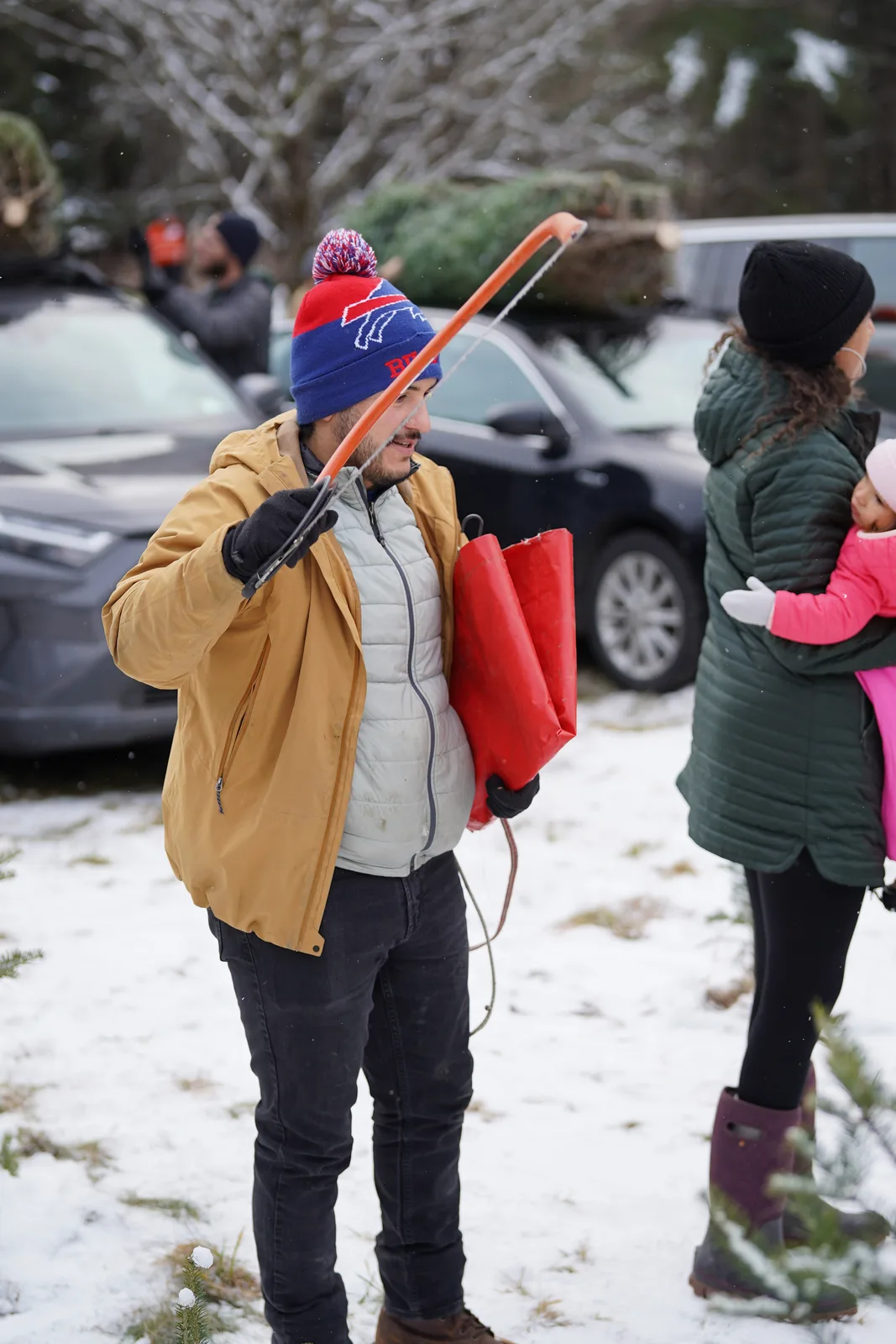 Man in a blue and red beanie holding a red saw and gas can in a snowy lot.