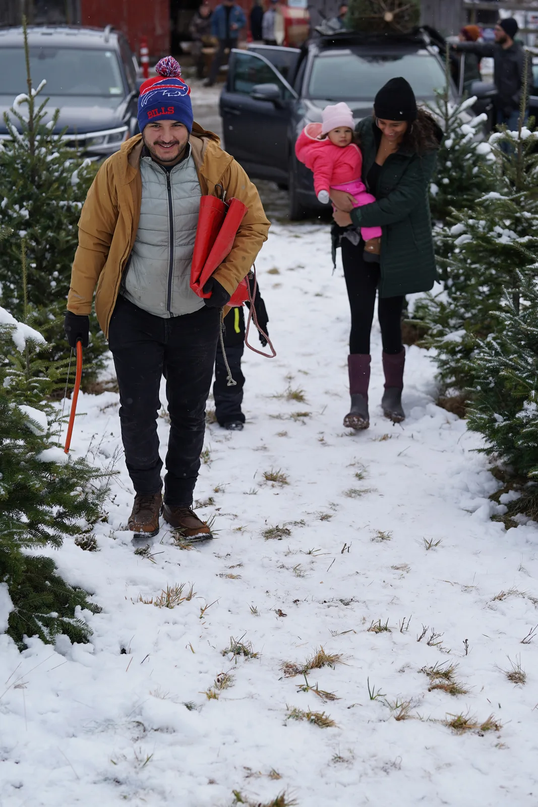 Family walking a snowy path between Christmas trees, one carrying a child in pink.