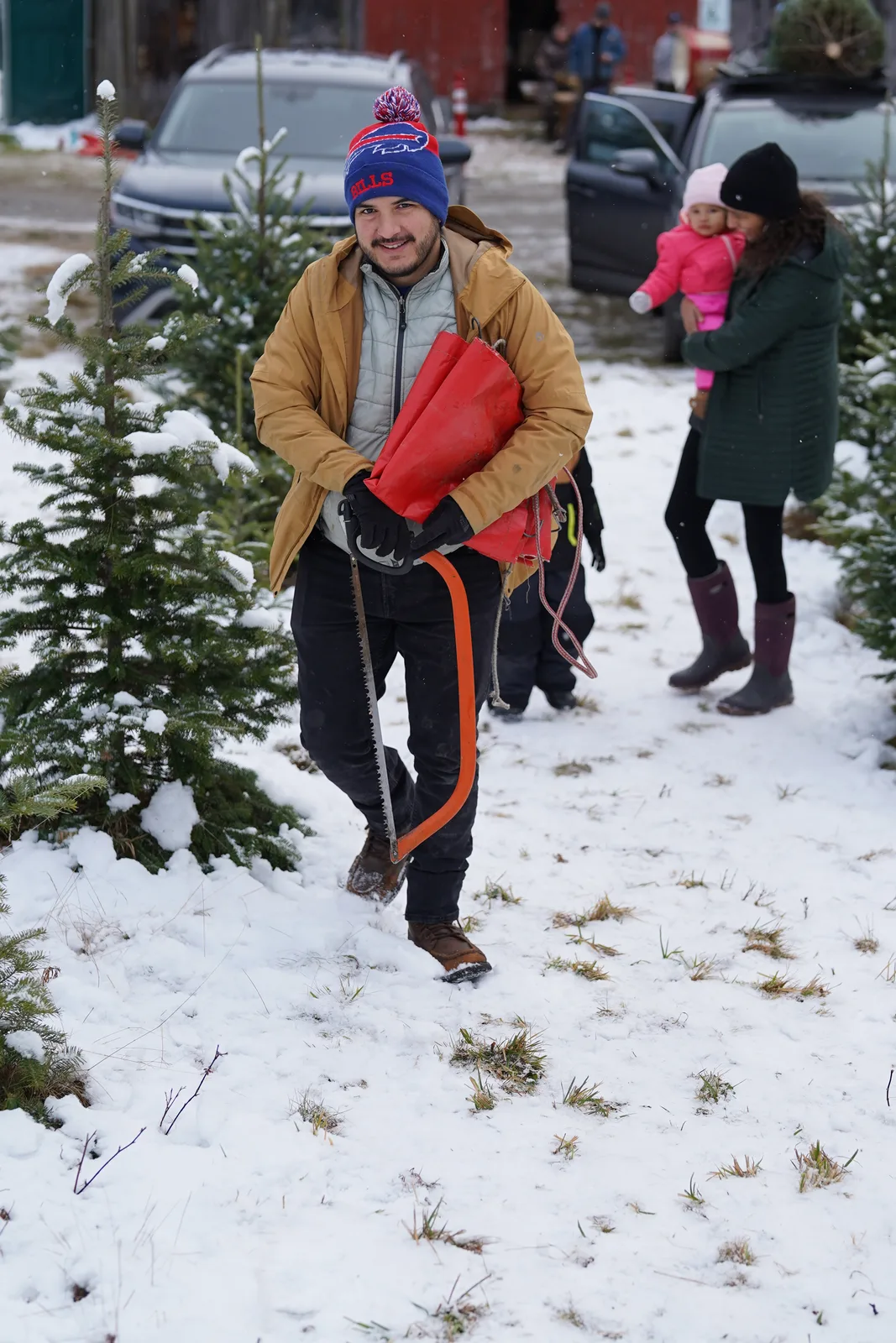 Man in winter gear carrying a red saw walks through snow among Christmas trees, two others in background