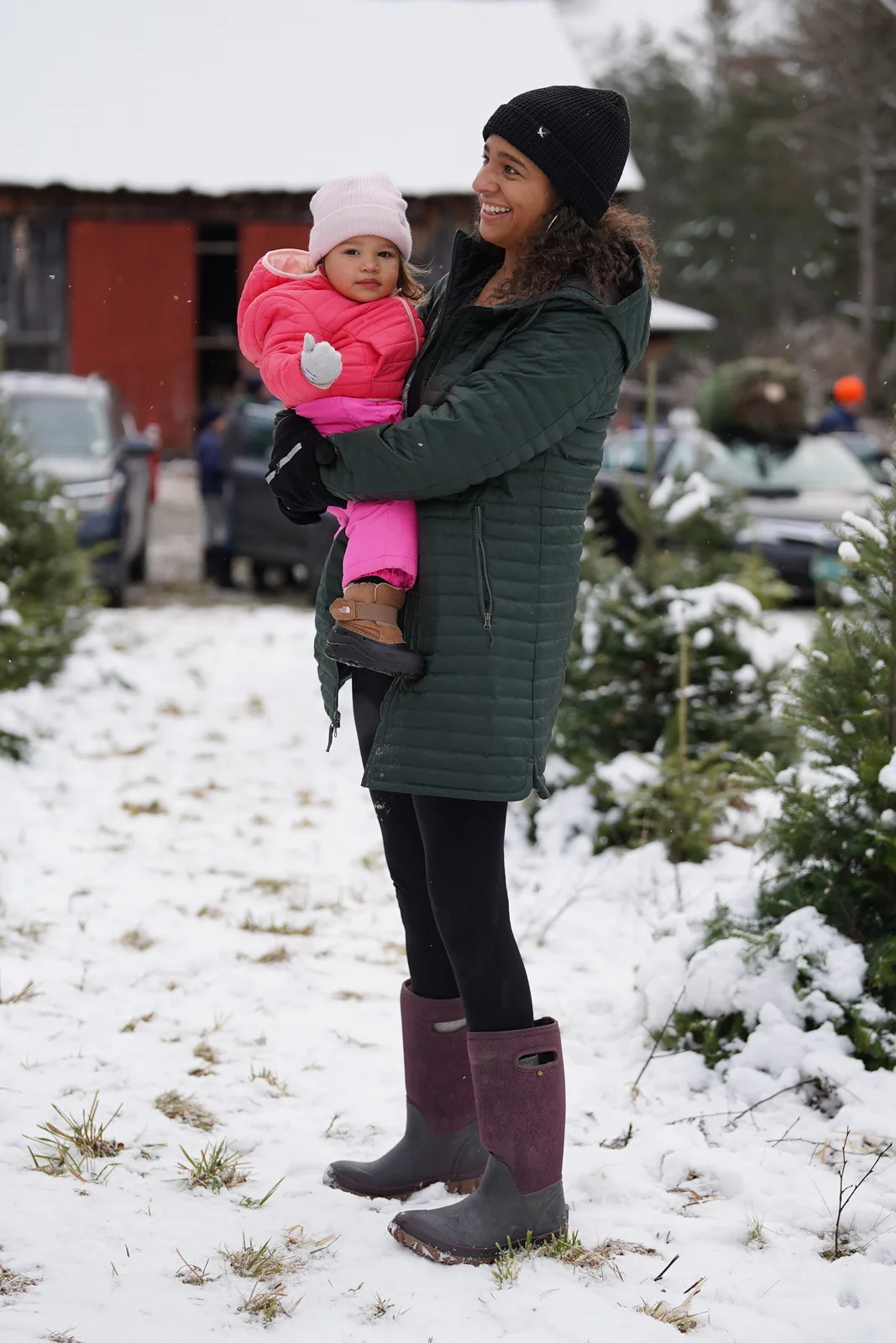 Woman in green coat and boots holds a child in pink snowsuit on snowy path