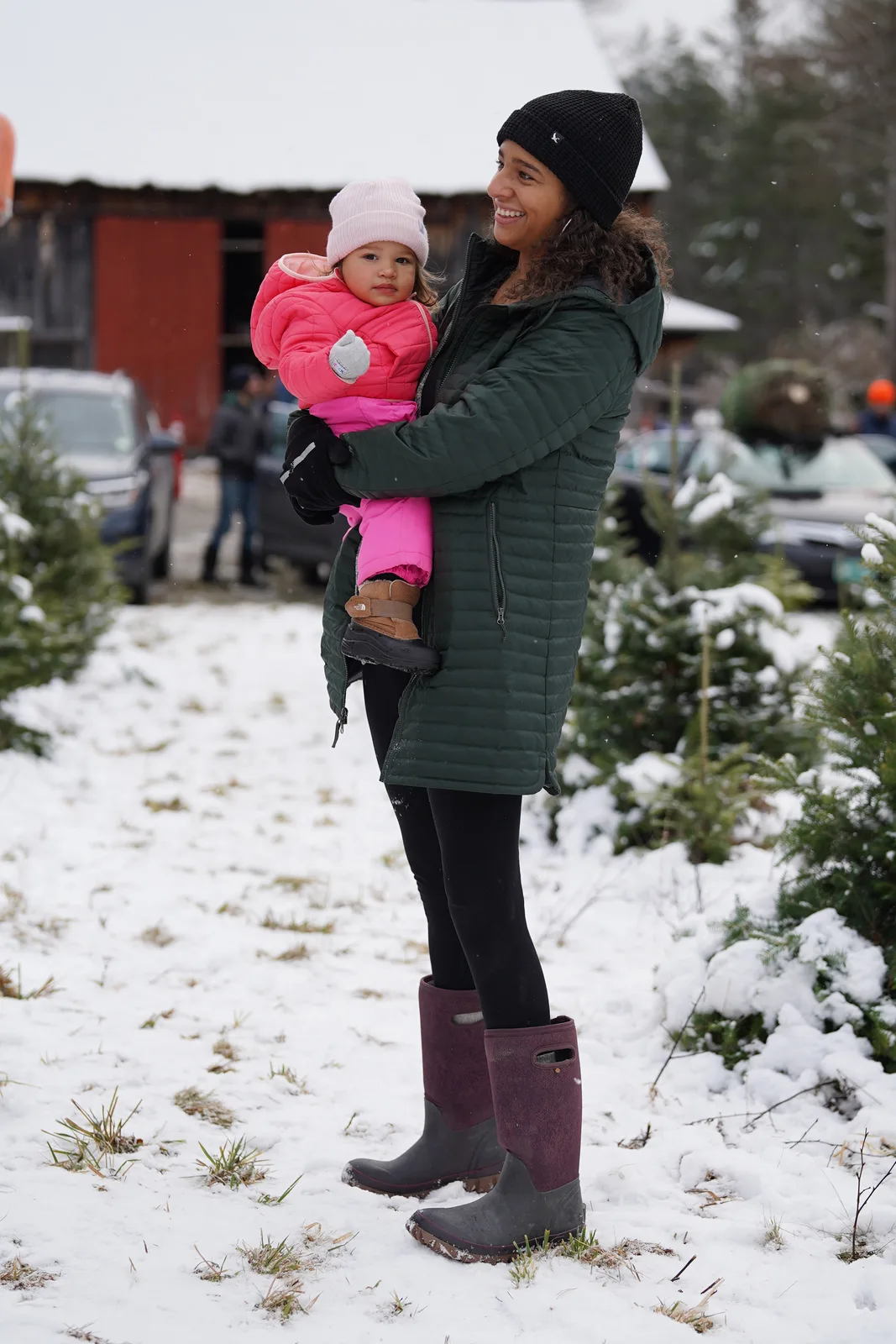 Woman in green coat and boots holds a child in pink snowsuit near snow-covered trees