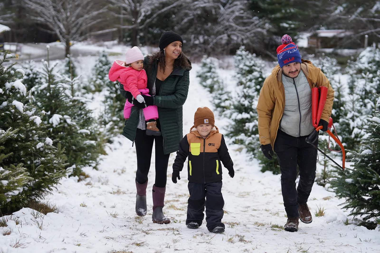 Four in winter coats on snowy path, child in pink held, red sleds carried, trees on both sides