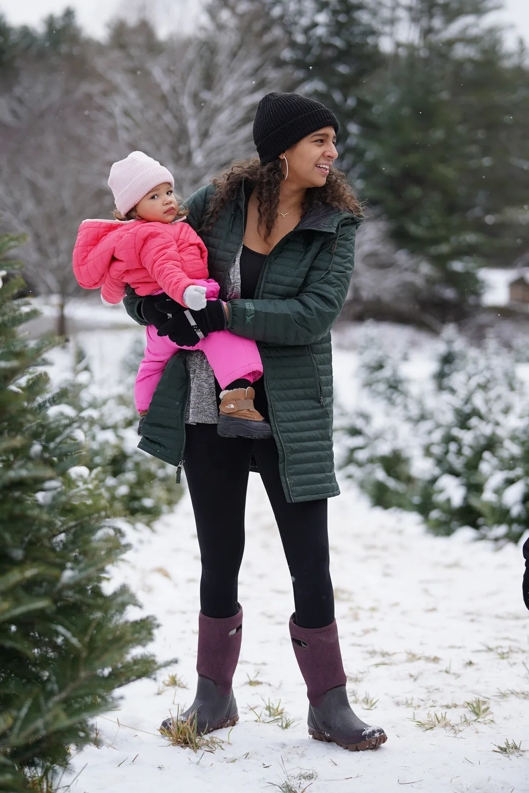 Woman in green coat and boots holds a child in pink snowsuit near snow-covered trees