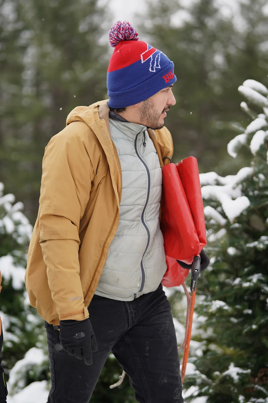 Person in mustard jacket, blue beanie with pom-pom, carrying red sled among snow-covered trees