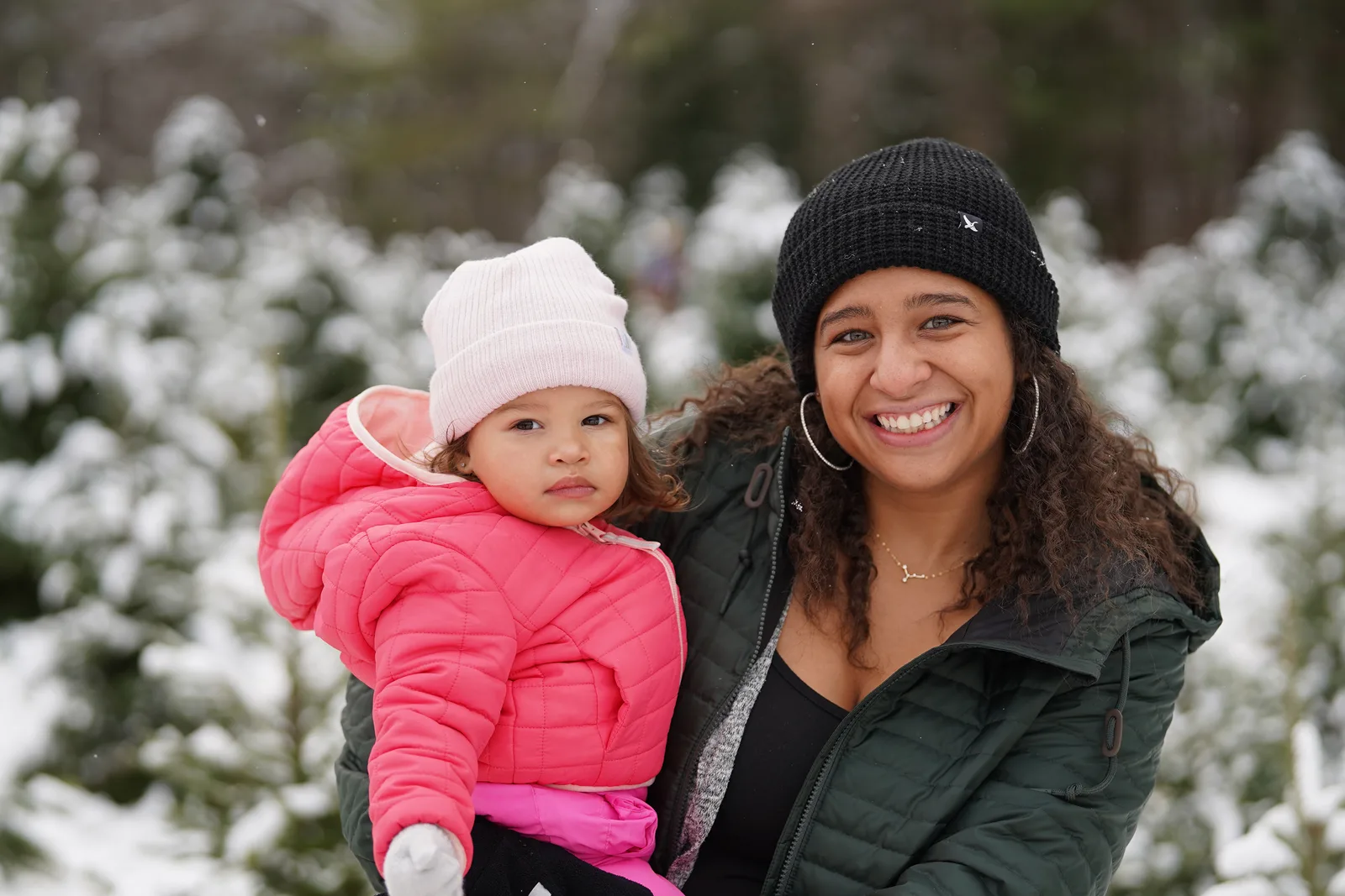Two people in winter coats, one in pink, one in green, among snow-dusted Christmas trees