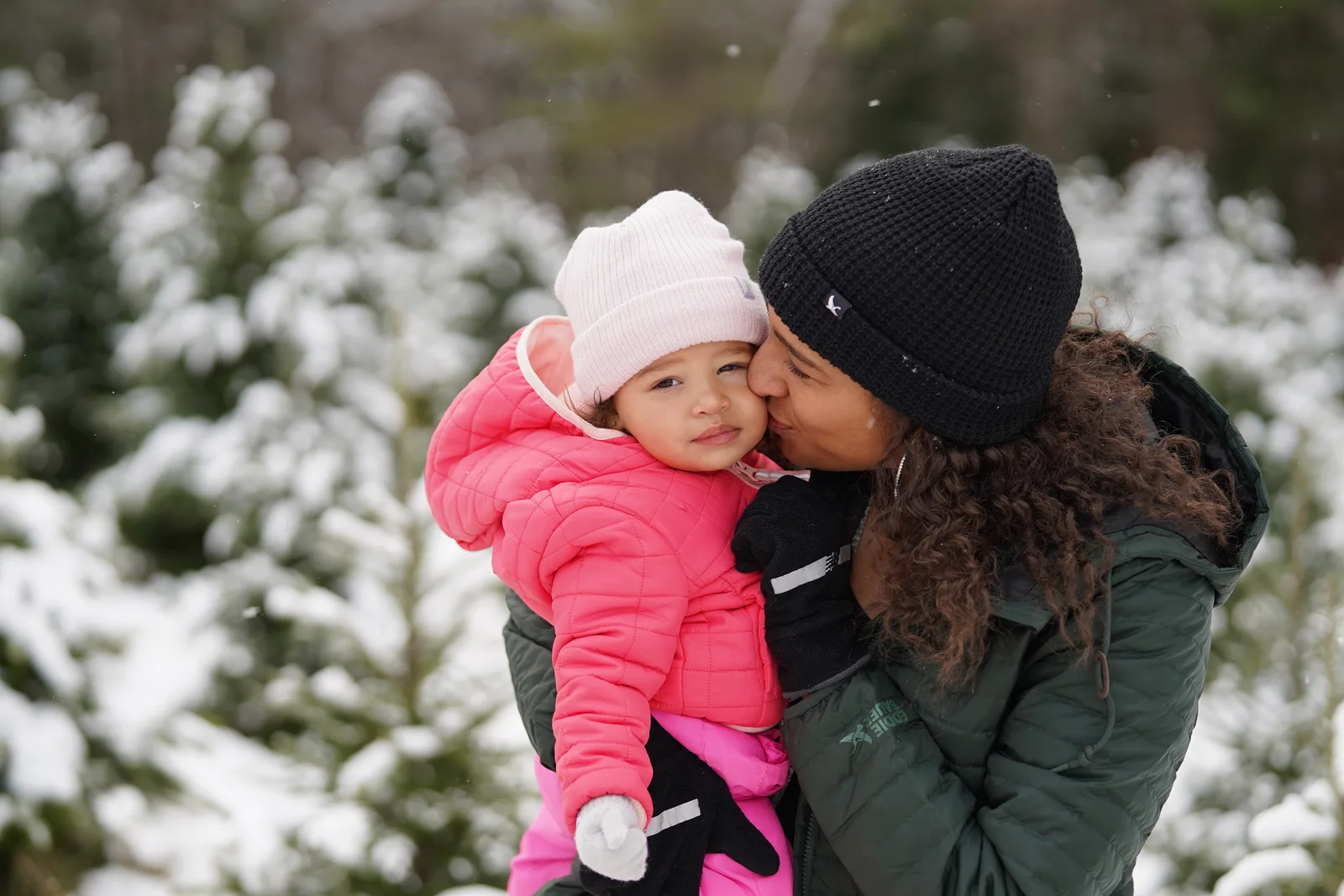 Person in green jacket kissing child in pink coat and light hat amid snow