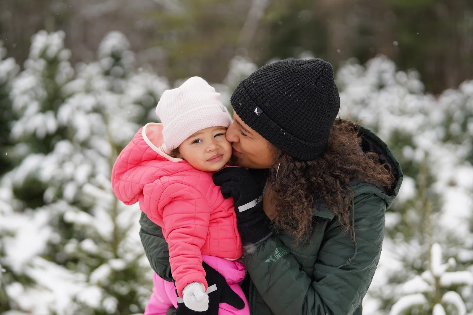 Person in green jacket kissing child in pink coat and light hat by snow-covered trees