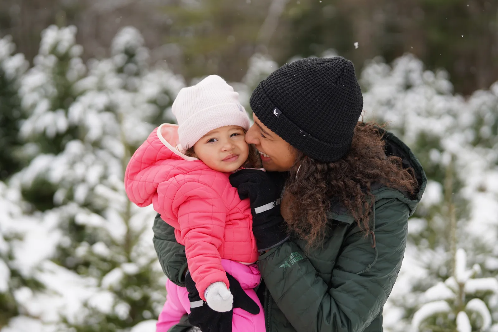 Person in green jacket holding child in pink coat and light hat near snowy trees