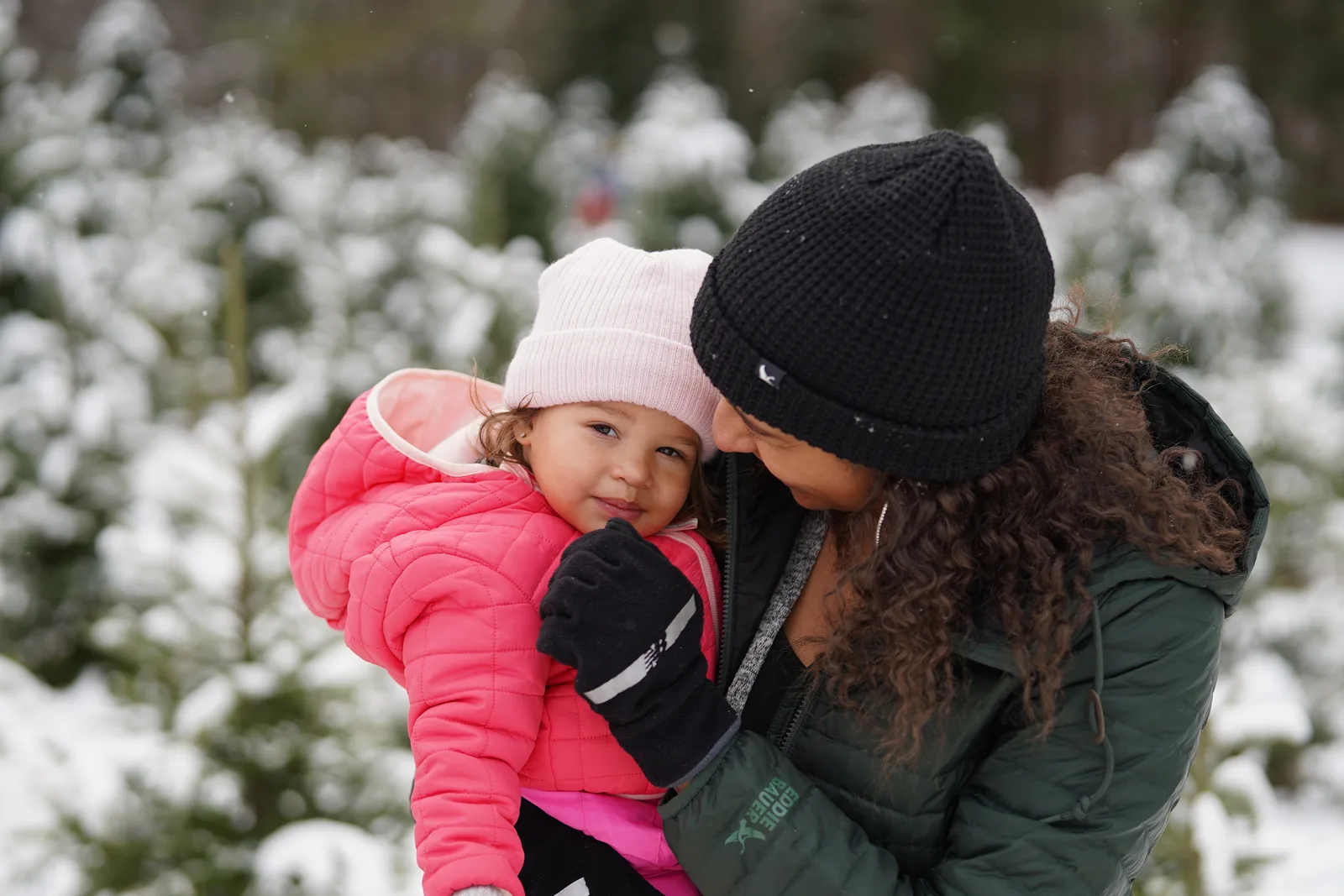 Person in green jacket and black beanie kissing child in pink coat and light hat