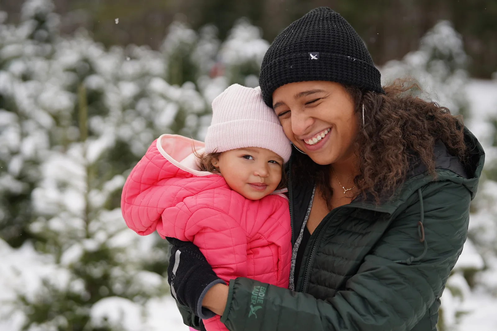 Adult in black beanie holding child in pink jacket and light hat among snow-covered trees