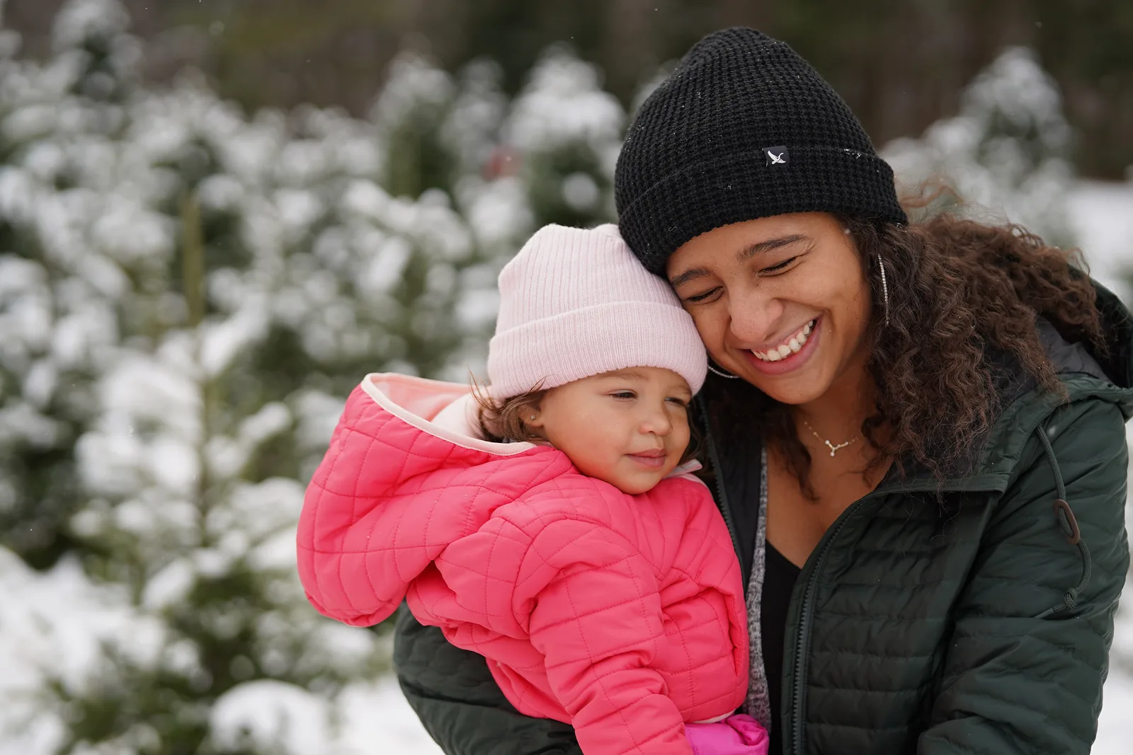 Adult in black beanie holding child in pink jacket and light hat by snow-dusted Christmas trees