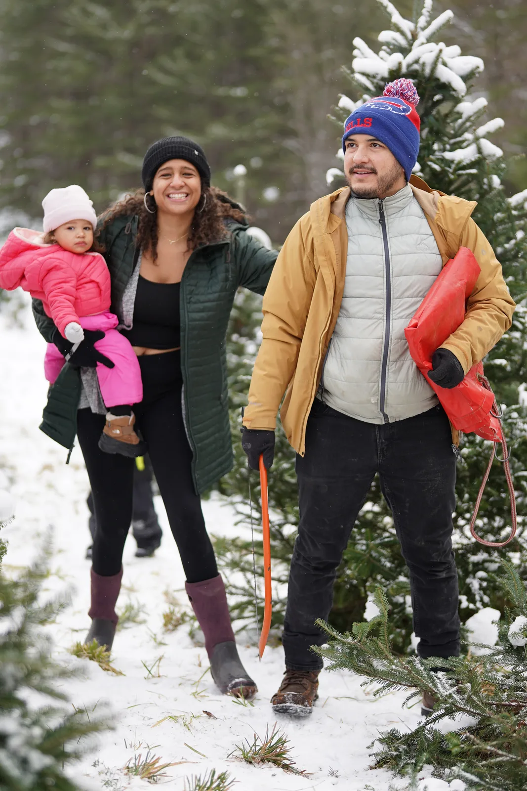 Three people in winter coats, one in pink, one in green, one in mustard, among snow-dusted Christmas trees