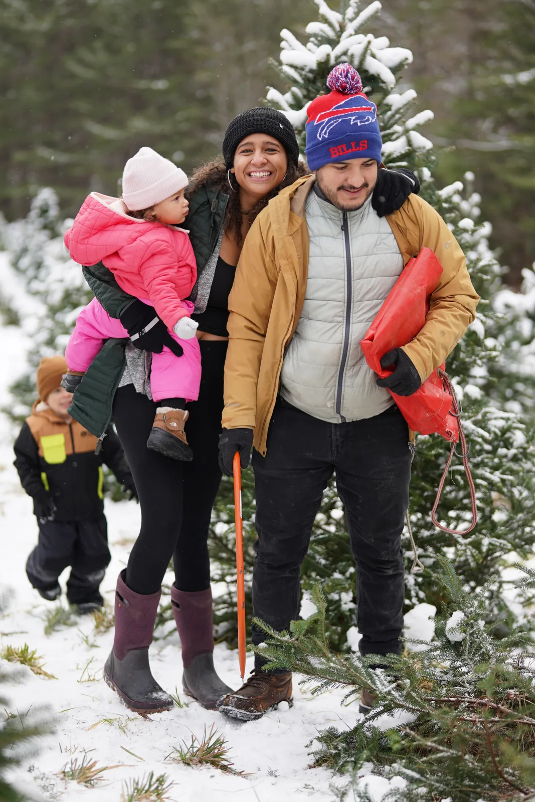 Four people in winter coats, one holding a child in pink, among snow-covered Christmas trees.