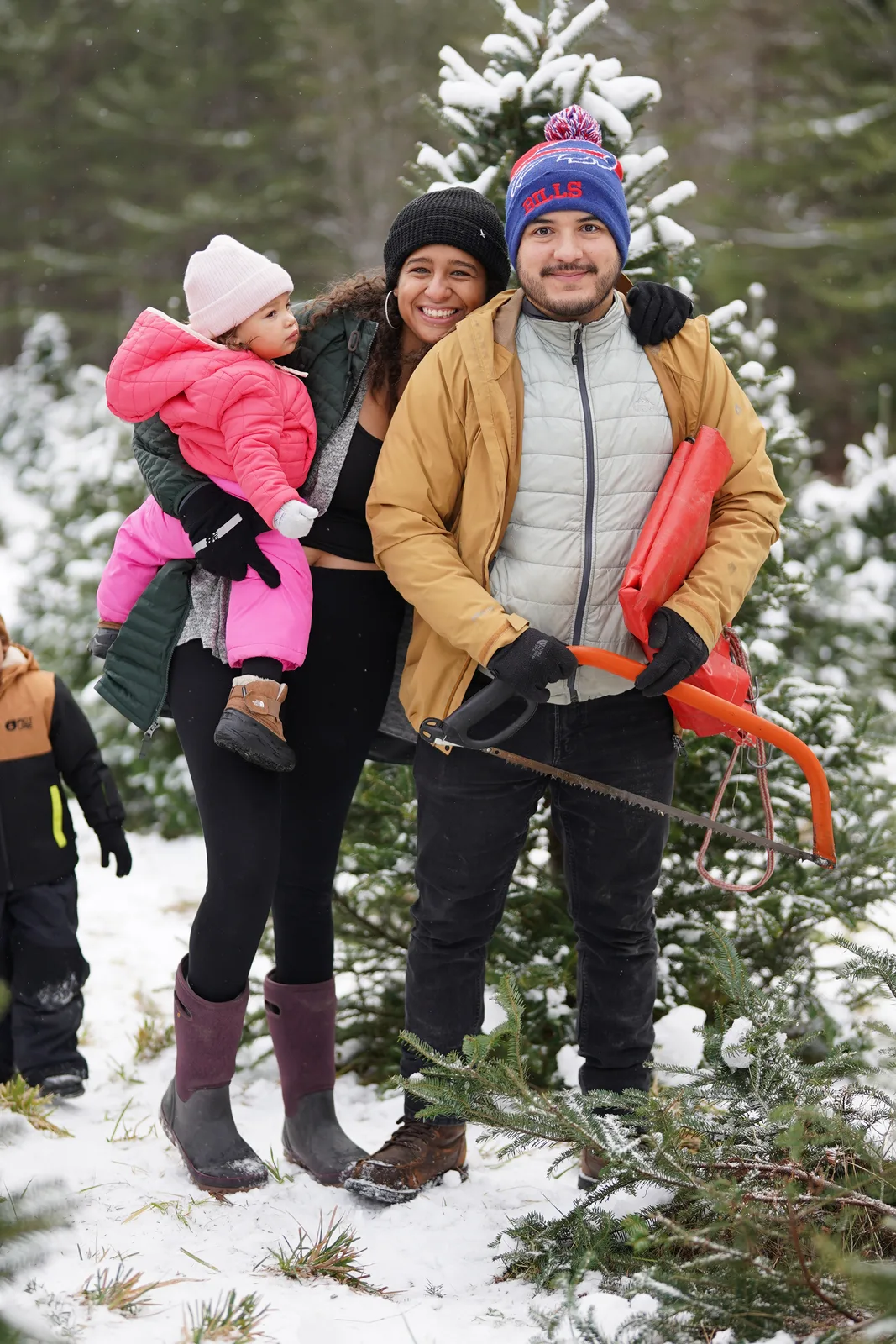 Three adults and a child in pink jacket stand among snow-dusted Christmas trees.