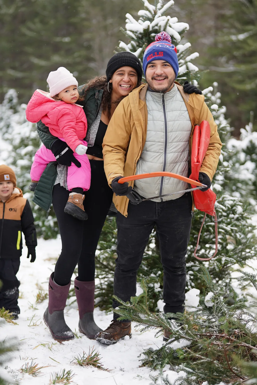 Three adults and a child in pink jacket stand among snow-laden Christmas trees.