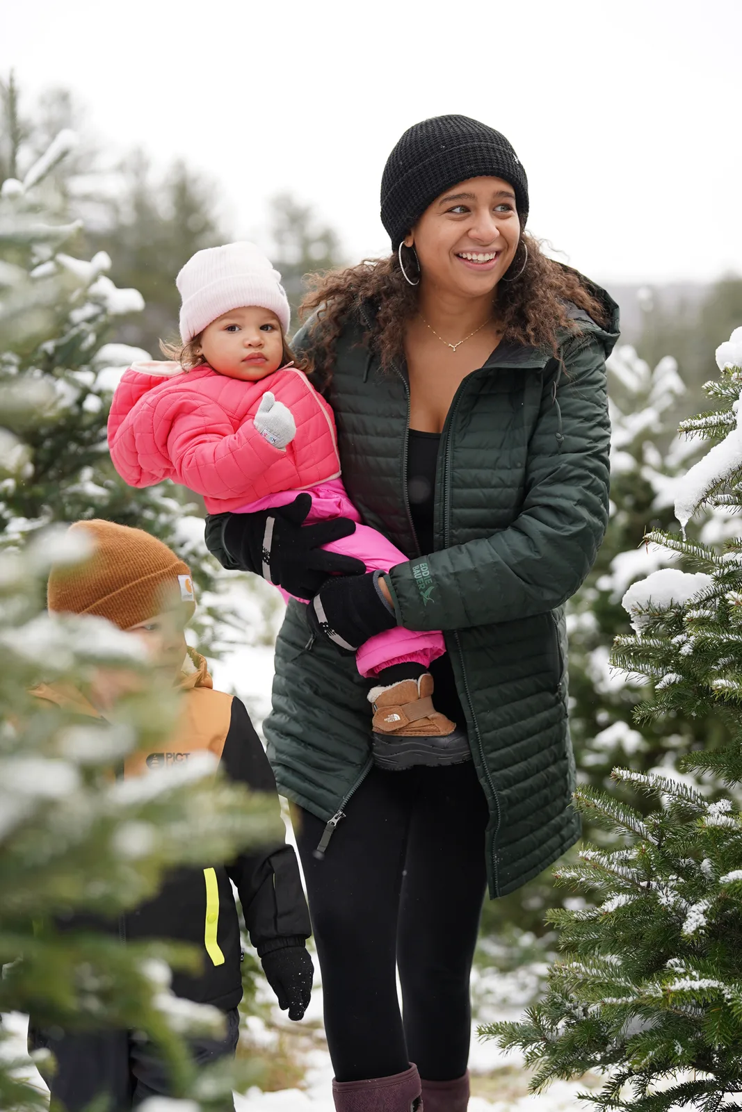 Woman in green jacket holds a child in pink coat among snow-covered trees.