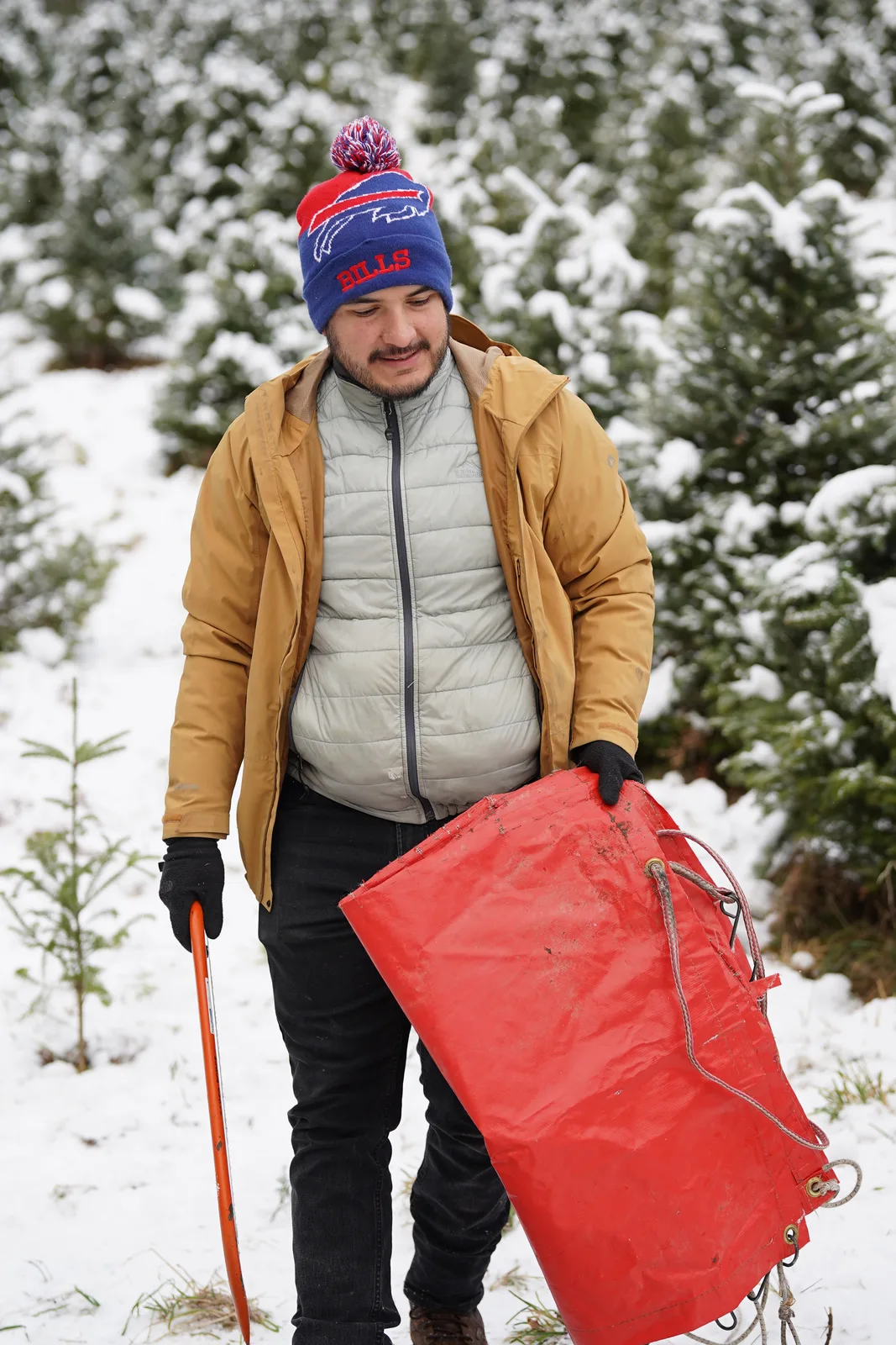Man in blue beanie and tan jacket carries red sled among snow-covered trees.