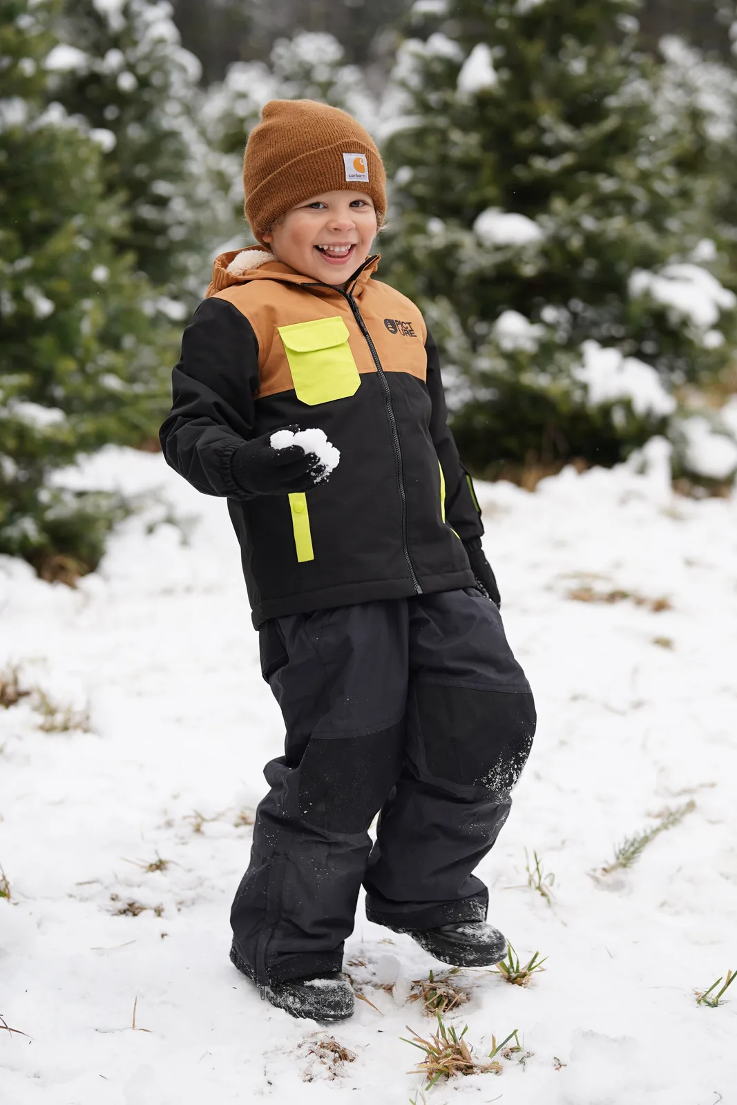 Child in brown beanie and black jacket holds snowball in snowy tree farm.