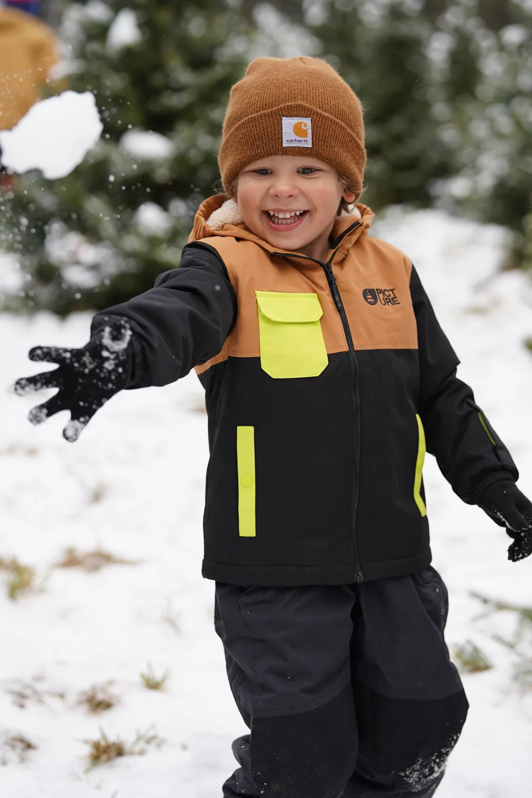 Child in brown beanie and black jacket with yellow accents reaching for snowflakes in a snowy tree farm.