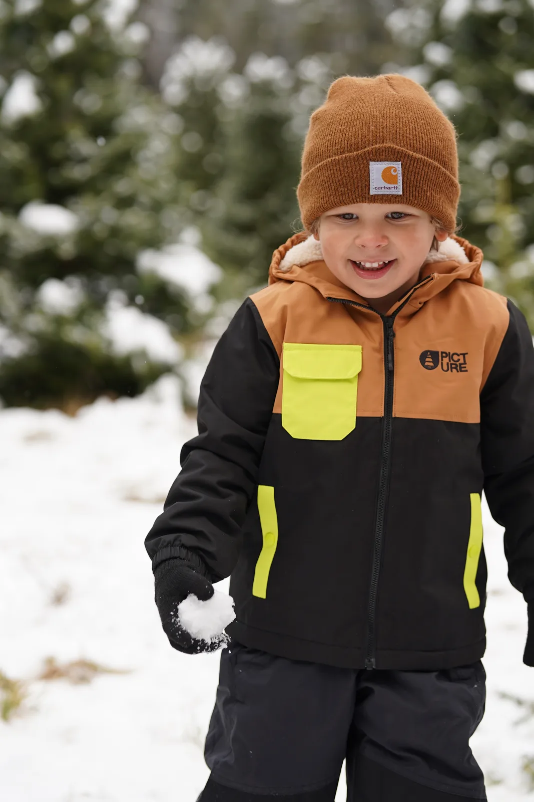Child in brown beanie and black jacket with yellow accents holding snowball among snow-covered trees.