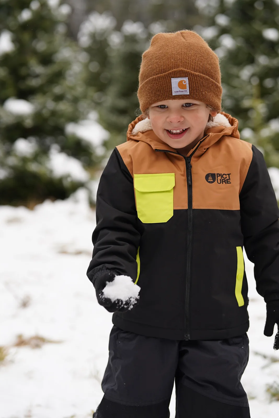 Child in brown beanie and black jacket with yellow accents holding snowball near evergreen trees.