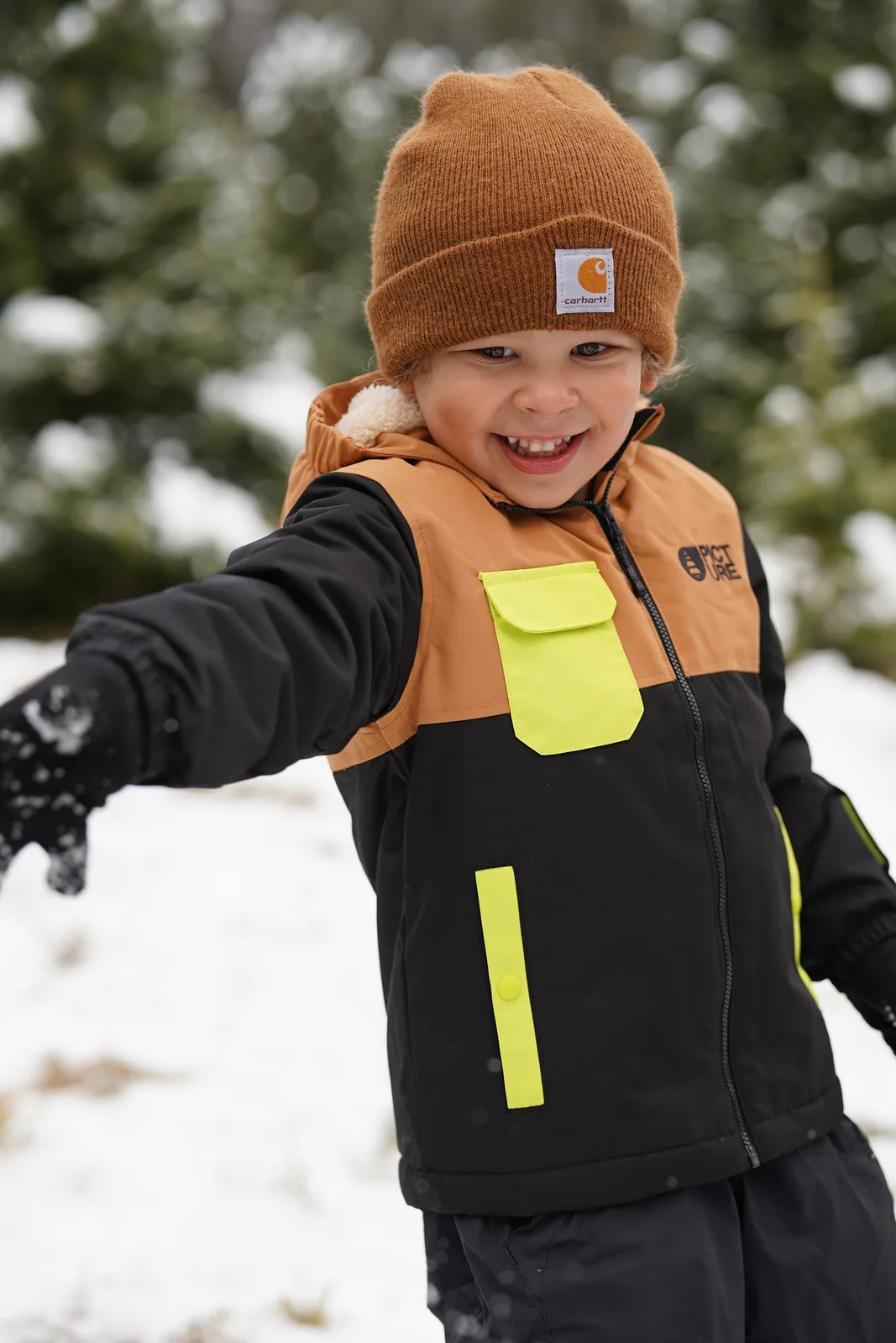 Child in brown beanie and black jacket with yellow accents reaching out in snowy tree farm.