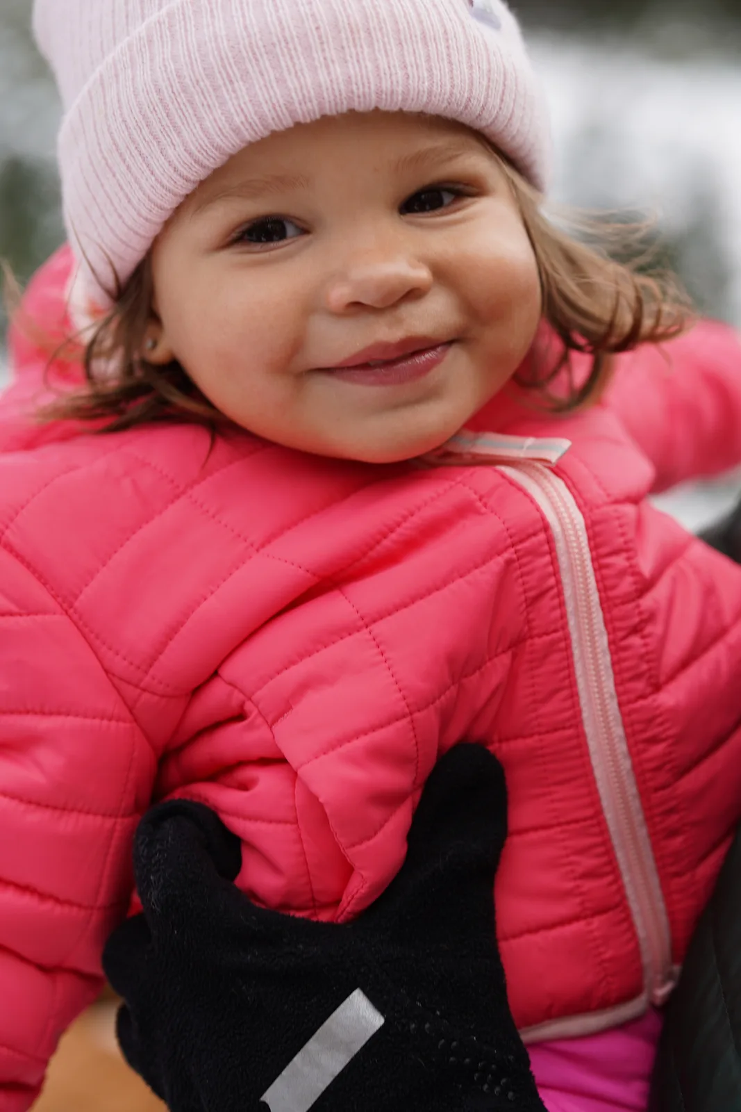 Child in pink beanie and pink quilted jacket with black gloves in snowy outdoor setting.
