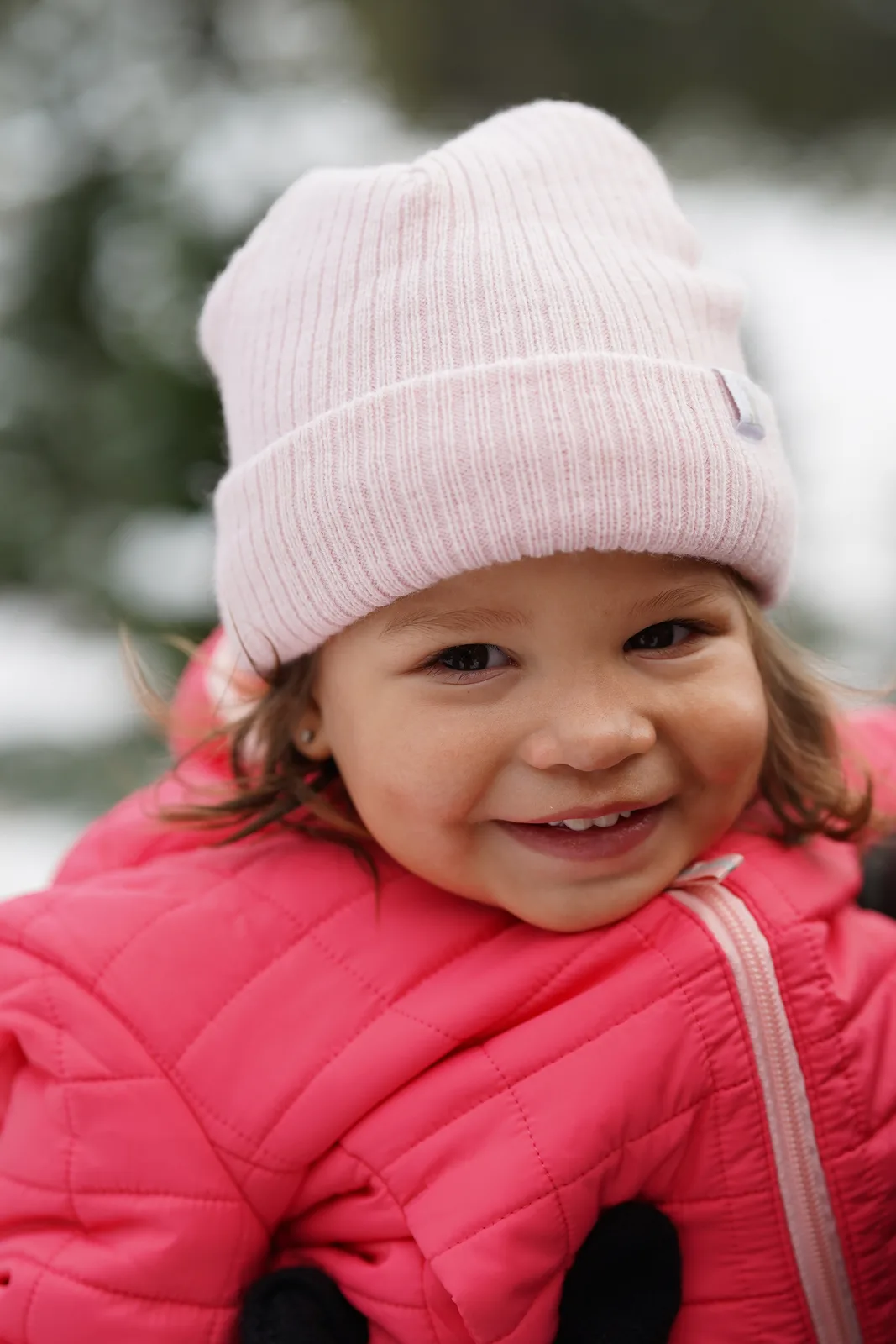 Child in pink beanie and pink quilted jacket smiling amid snow-dusted Christmas trees.