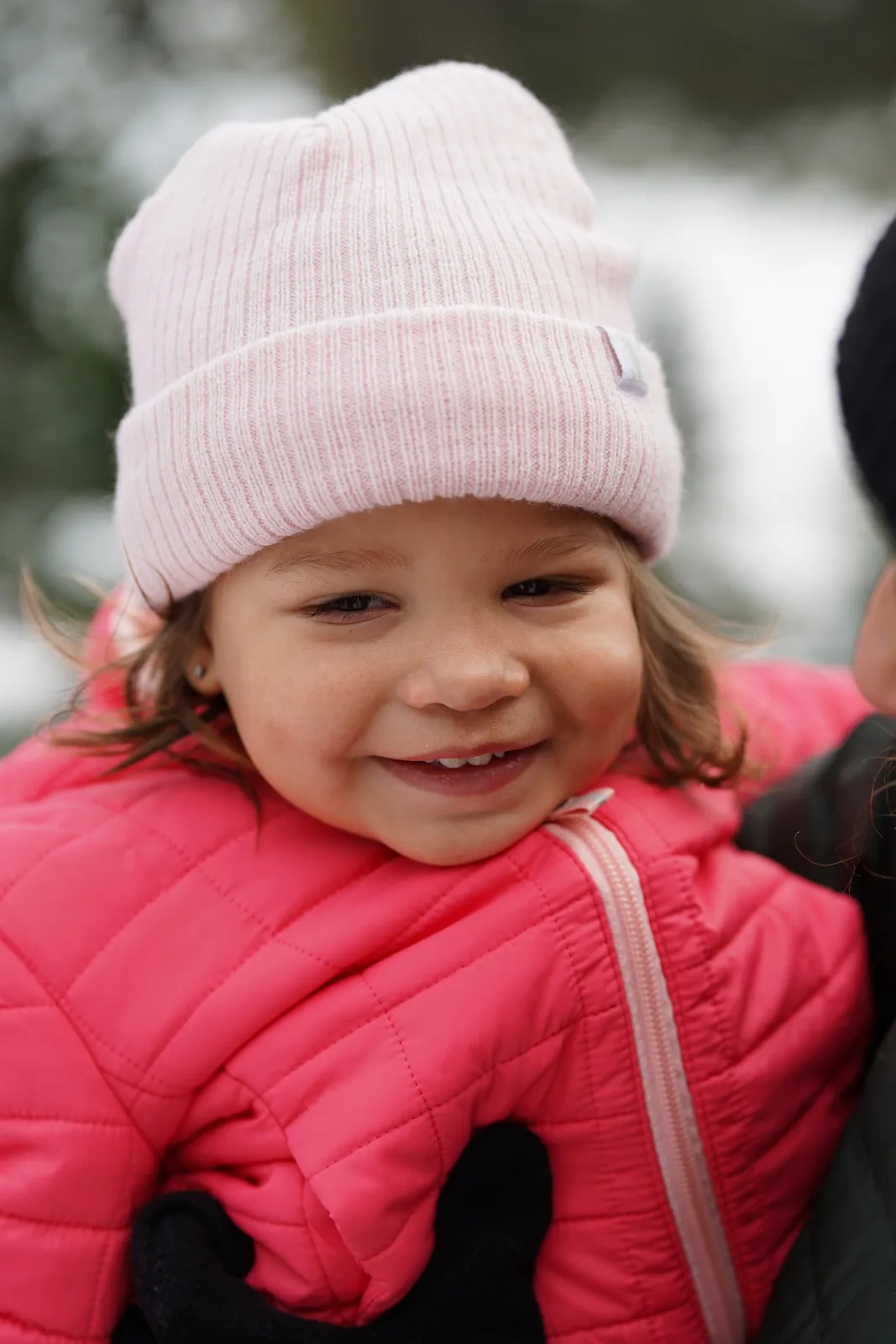 Child in pink beanie and pink quilted jacket smiling near snow-covered evergreen trees.