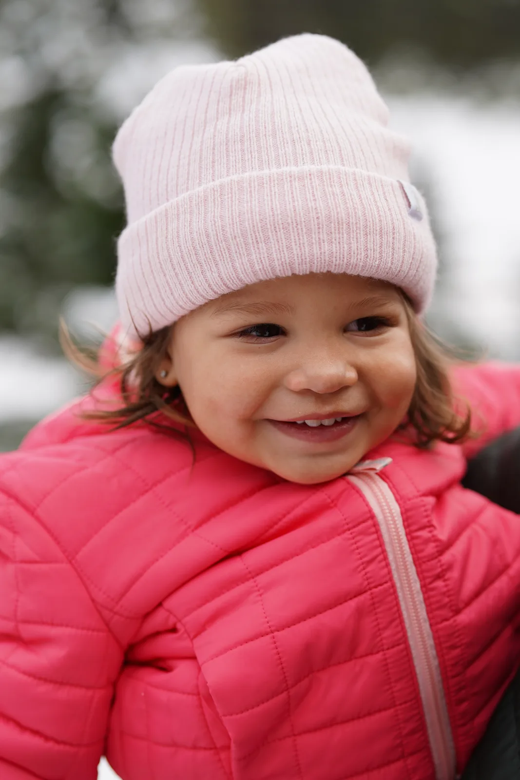 Child in pink beanie and pink quilted jacket smiling by snowy Christmas trees.
