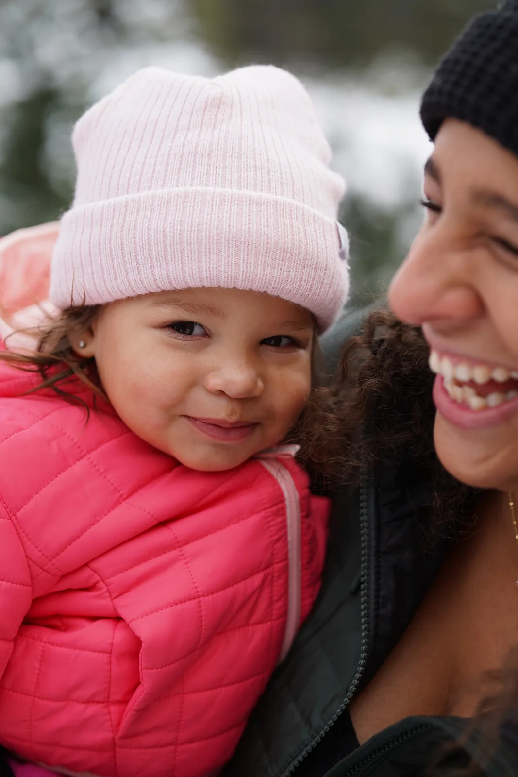 Child in pink beanie and pink quilted jacket held by adult in dark coat among snow-dusted trees