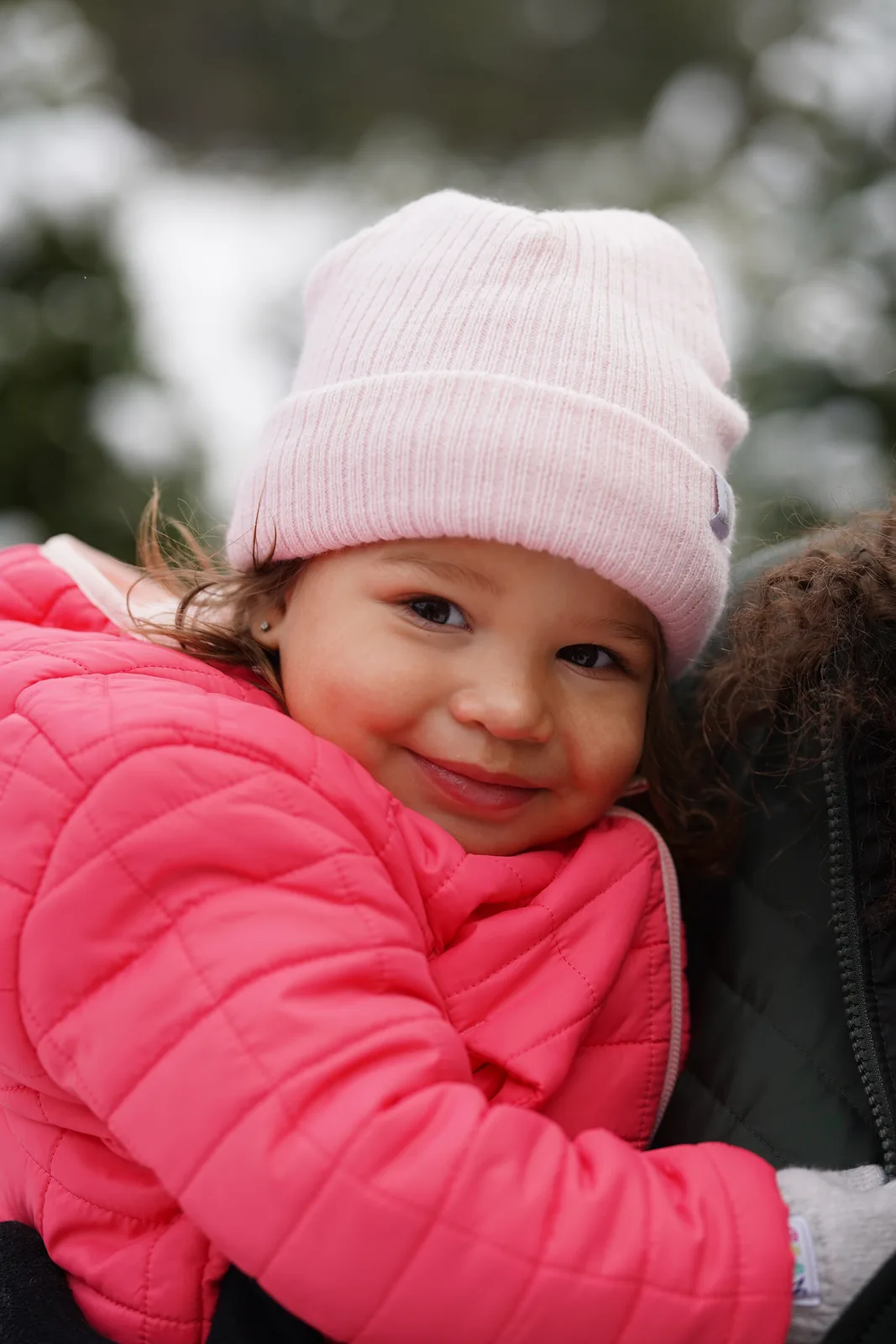 Child in pink beanie and pink jacket smiling while held by adult in dark coat