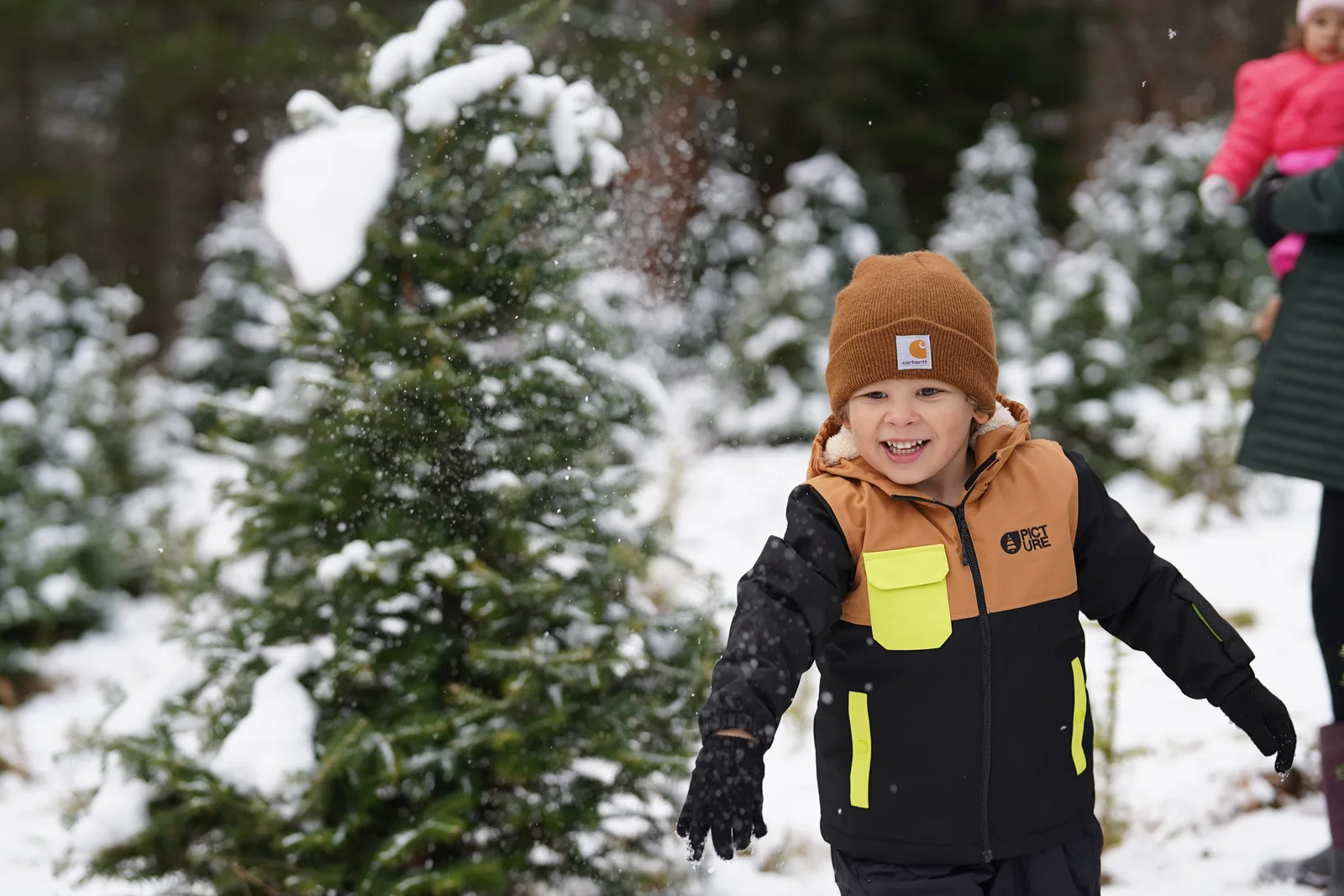 Child in brown beanie and black jacket with yellow accents throwing snow near evergreen trees