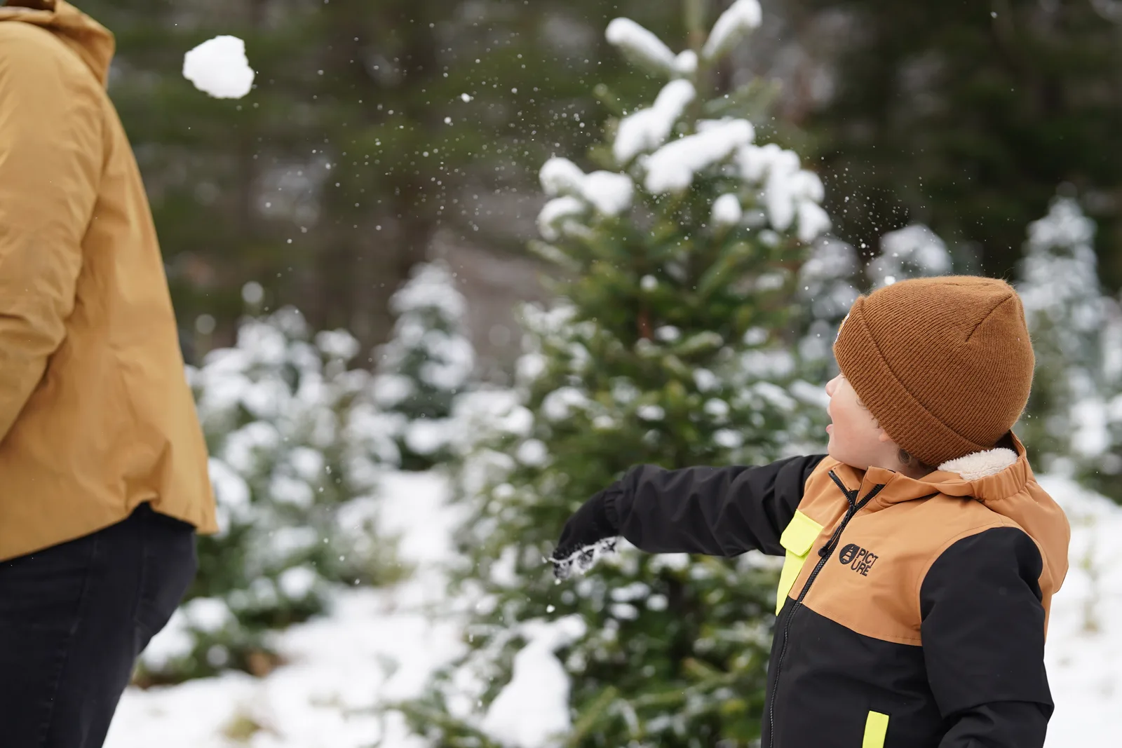Child in brown beanie and black jacket with yellow accents throwing snow at tree