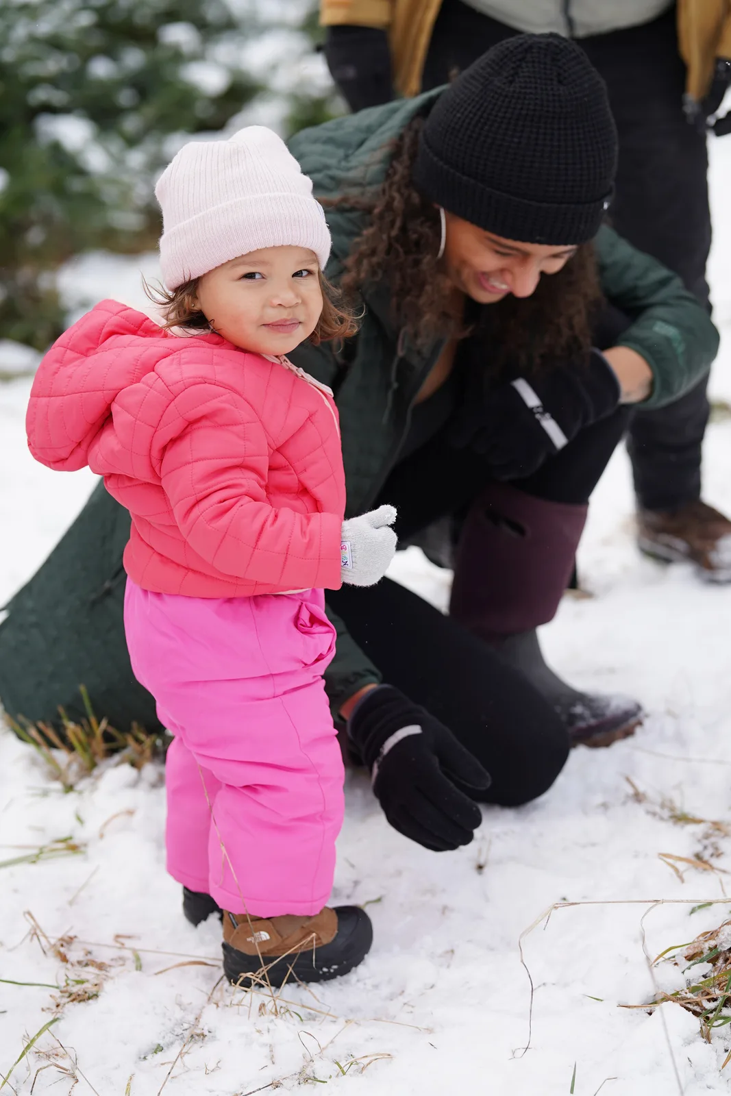 Child in pink beanie and pink snowsuit standing on snow near adult in black beanie