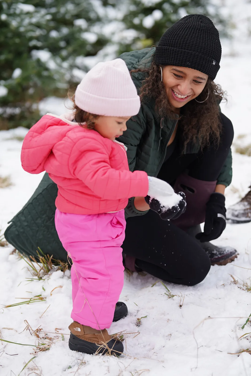 Child in pink beanie and pink snowsuit holding snowball with adult in black beanie