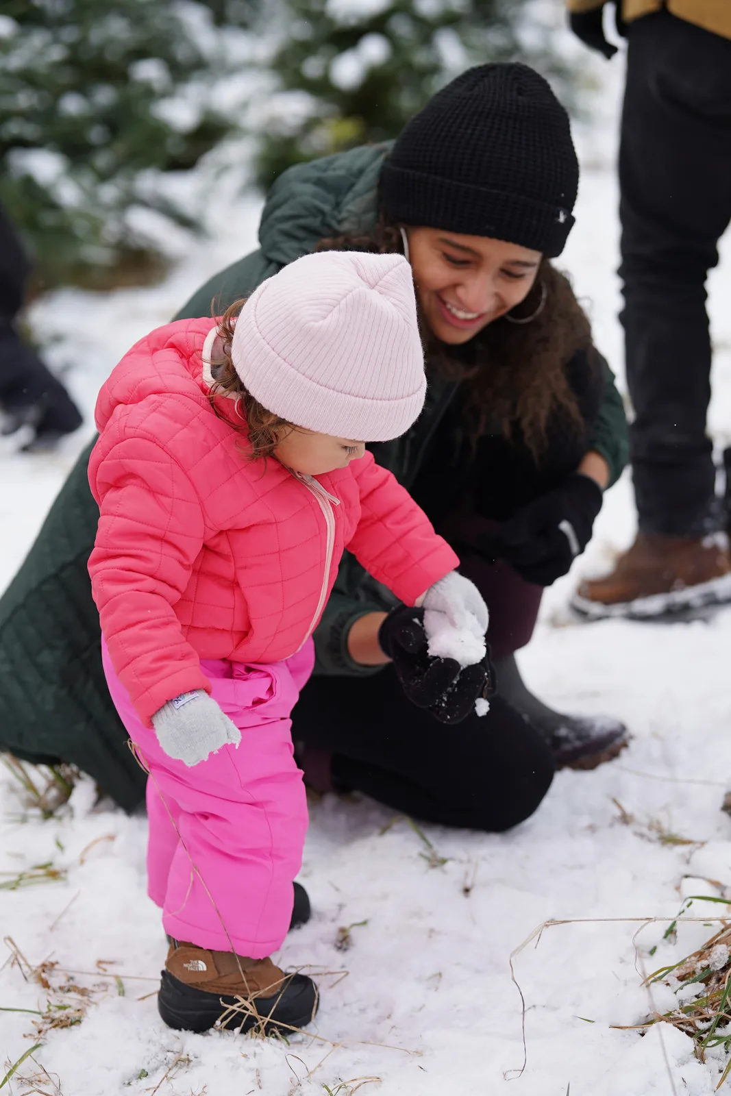 Child in pink beanie and pink snowsuit holding snowball with adult in black beanie