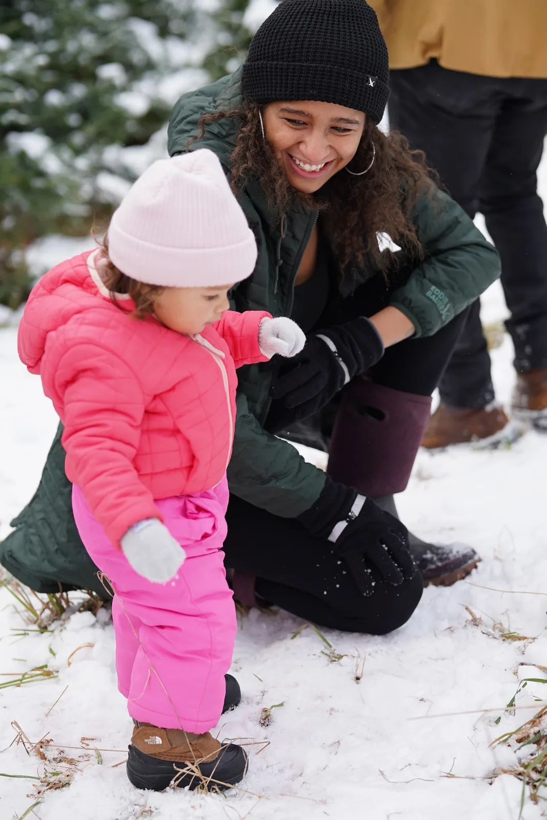 Adult in black beanie and green jacket crouching beside child in pink snowsuit and light hat on snow.