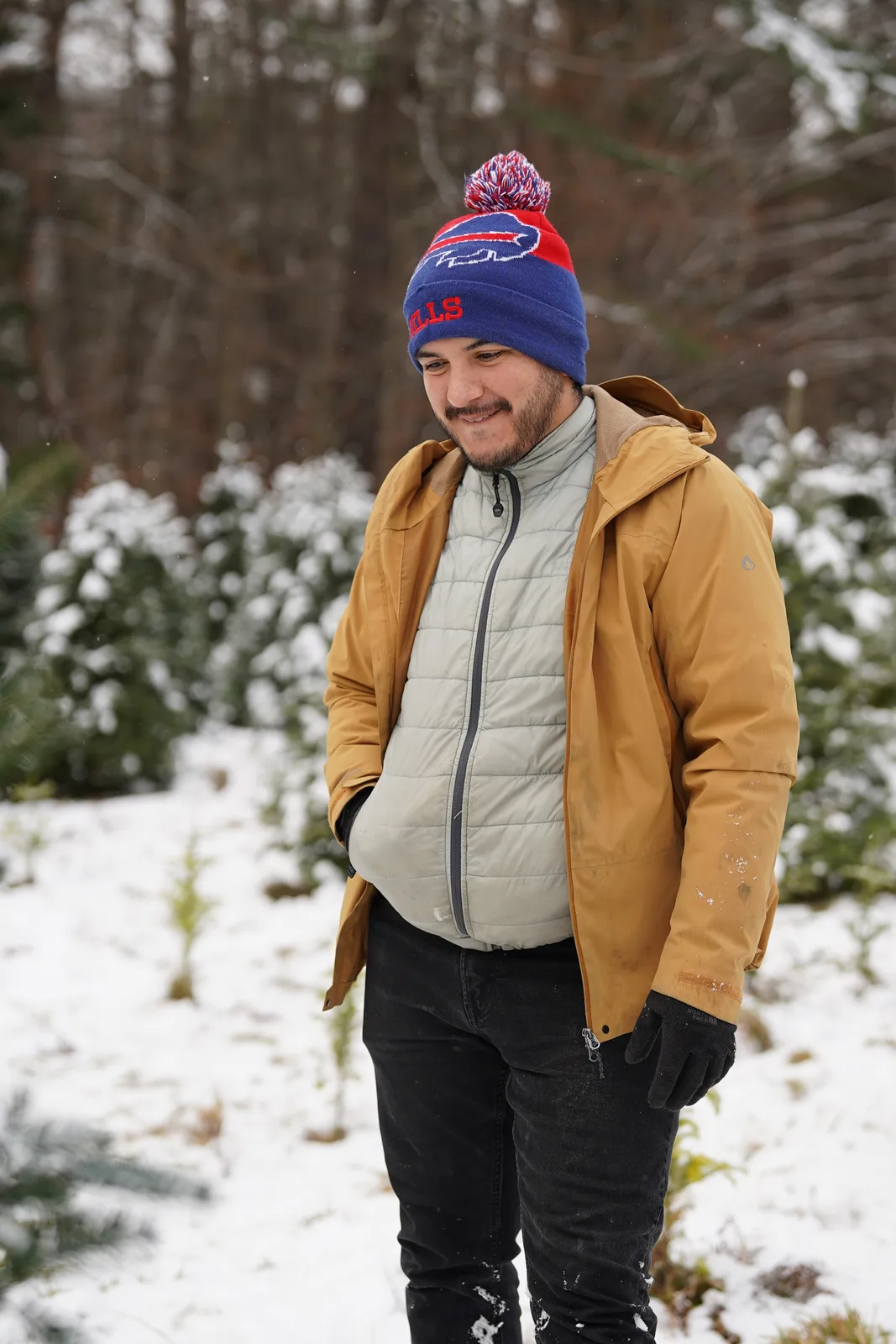 Person in tan jacket and blue beanie with pom-pom standing in snow among evergreen trees.