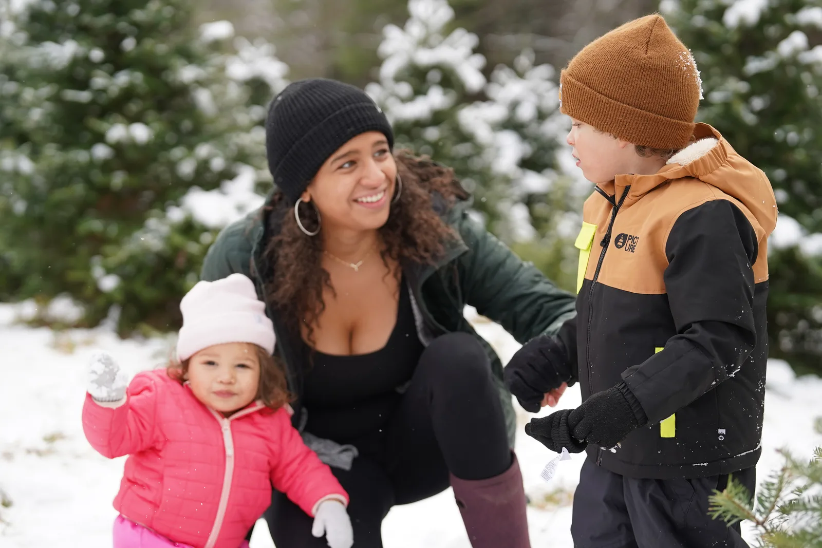 Adult in black beanie and green jacket with child in pink snowsuit and light hat beside child in brown beanie and black jacket.